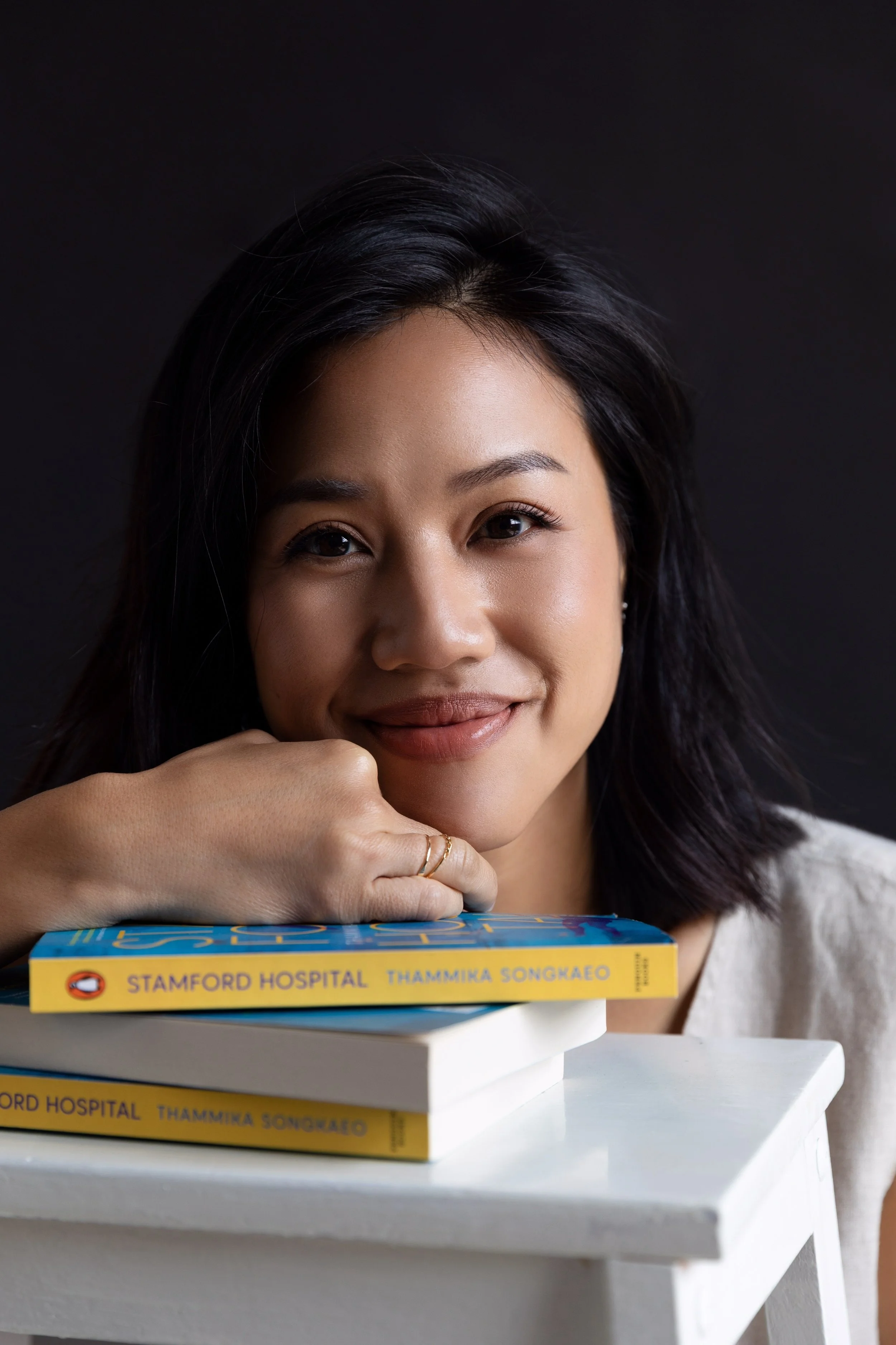 Close-up of a woman with black hair, wearing makeup, resting her chin on her hand, and smiling while looking at the camera. She is sitting behind a stack of books, with the top book titled 'Stamford Hospital' by Thammika Songkaeo, on a white table ag