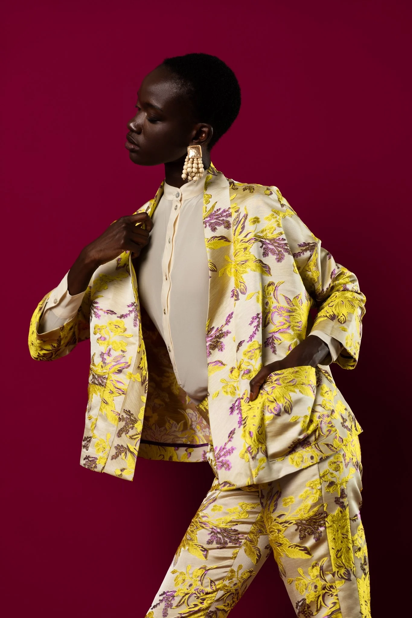 A woman with dark skin and short hair, dressed in a yellow and cream floral suit with purple accents, posing against a solid magenta background.
