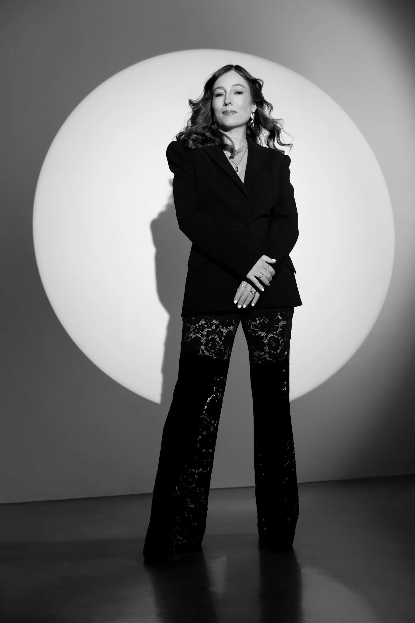 Black and white photo of a woman standing confidently in front of a spotlight circle. She has long, wavy hair, wears a blazer, patterned pants, and jewelry.