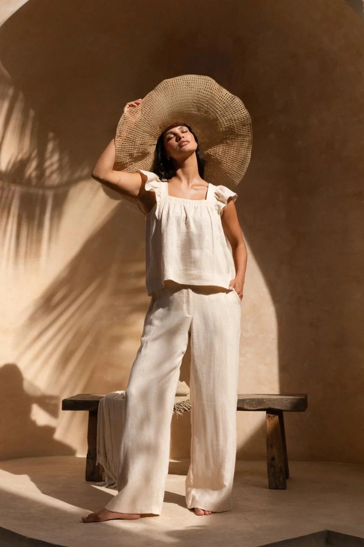 A woman wearing a wide-brimmed straw hat, cream-colored top, and matching loose-fitting pants, stands on a small wooden platform with one foot slightly raised, in a warm-toned room with textured beige walls. Shadows cast by the hat create patterns on