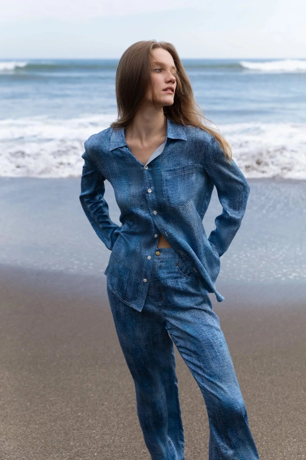 A woman with long, wavy brown hair wearing a denim jacket and jeans standing on the beach with waves in the background.