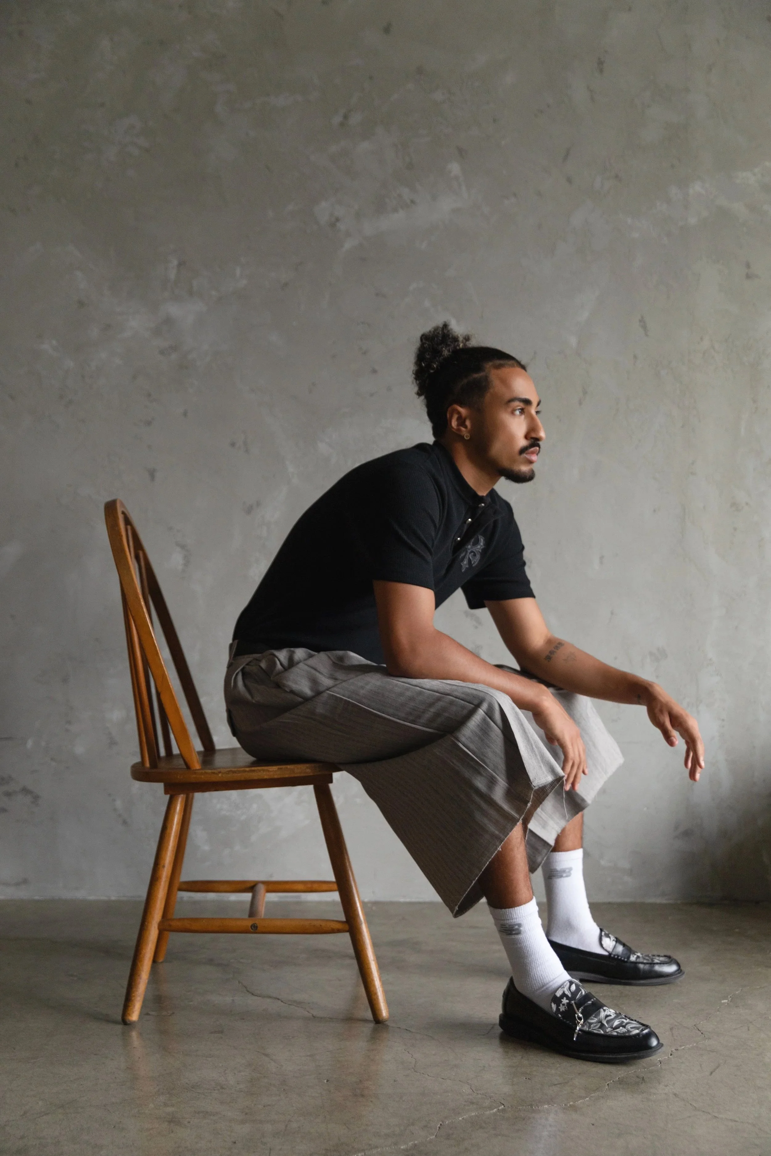 A young man sitting on a wooden chair against a gray textured wall, wearing a black shirt, plaid shorts, white socks, and patterned black shoes, with his hands resting on his knees and looking to the right.