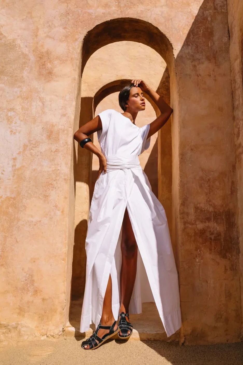 A woman in a white dress with high slits standing in an arched corridor with sand-colored walls.
