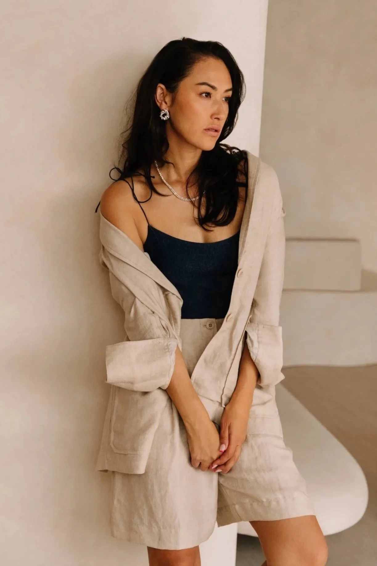 A woman with long dark hair wearing a black tank top, beige blazer, and beige shorts, accessorized with earrings and a necklace, standing against a plain wall.