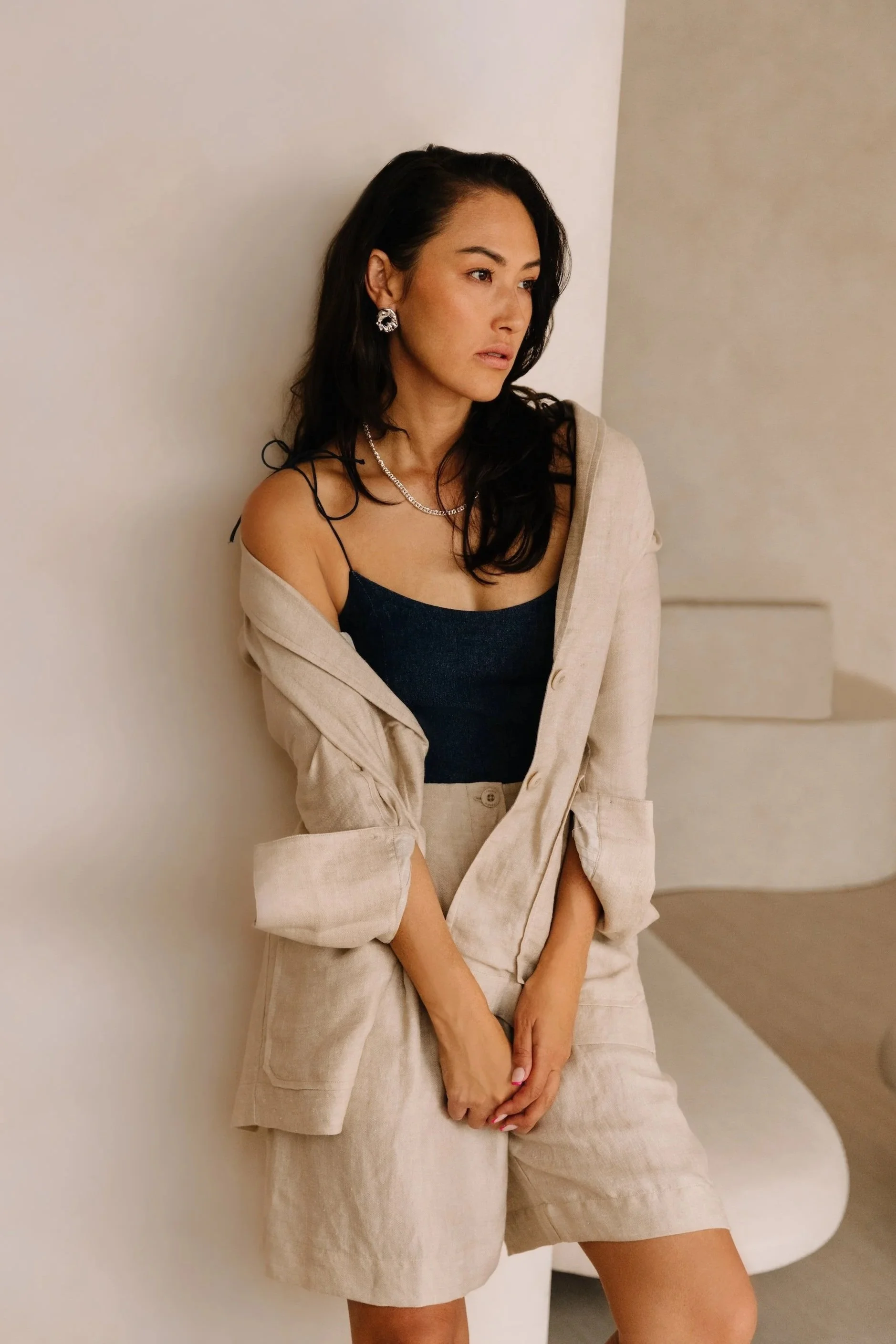 A woman with long dark hair and earrings wearing a black top, beige blazer, and beige shorts, standing against a white wall.