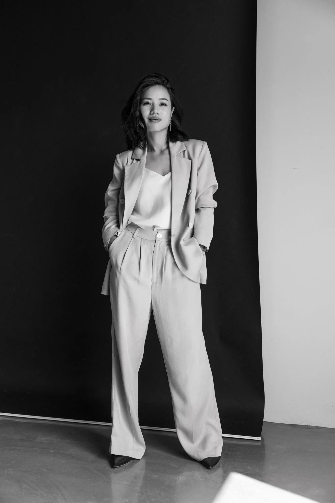 Black and white photo of a woman standing indoors, wearing a tailored blazer, high-waisted trousers, and pointed shoes. She has shoulder-length hair styled in waves and is posing confidently with one hand in her pocket, looking at the camera.