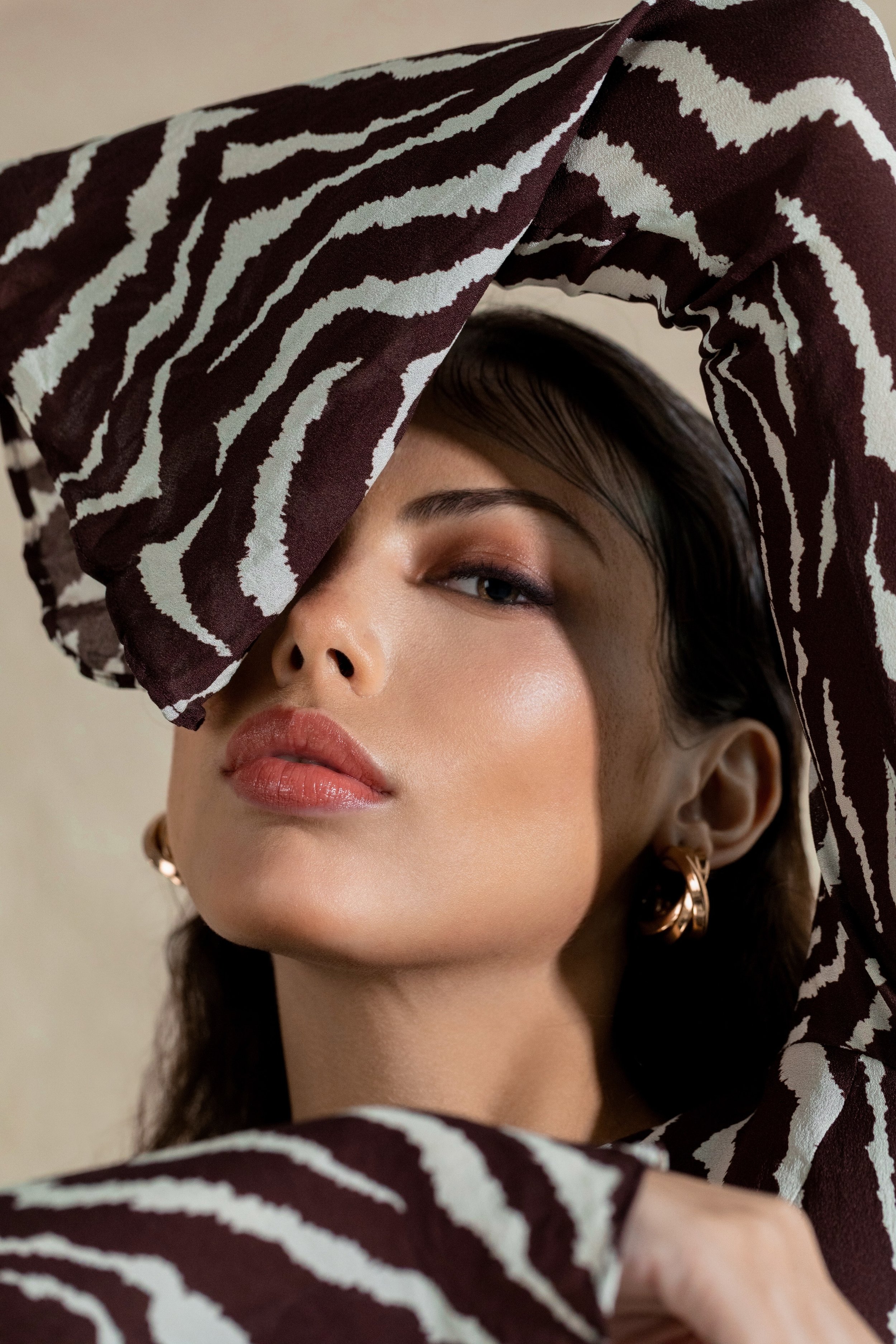 A woman with makeup and black hair, wearing gold hoop earrings, partially covered by a zebra-patterned scarf or fabric, with her face slightly tilted and one eye closed.