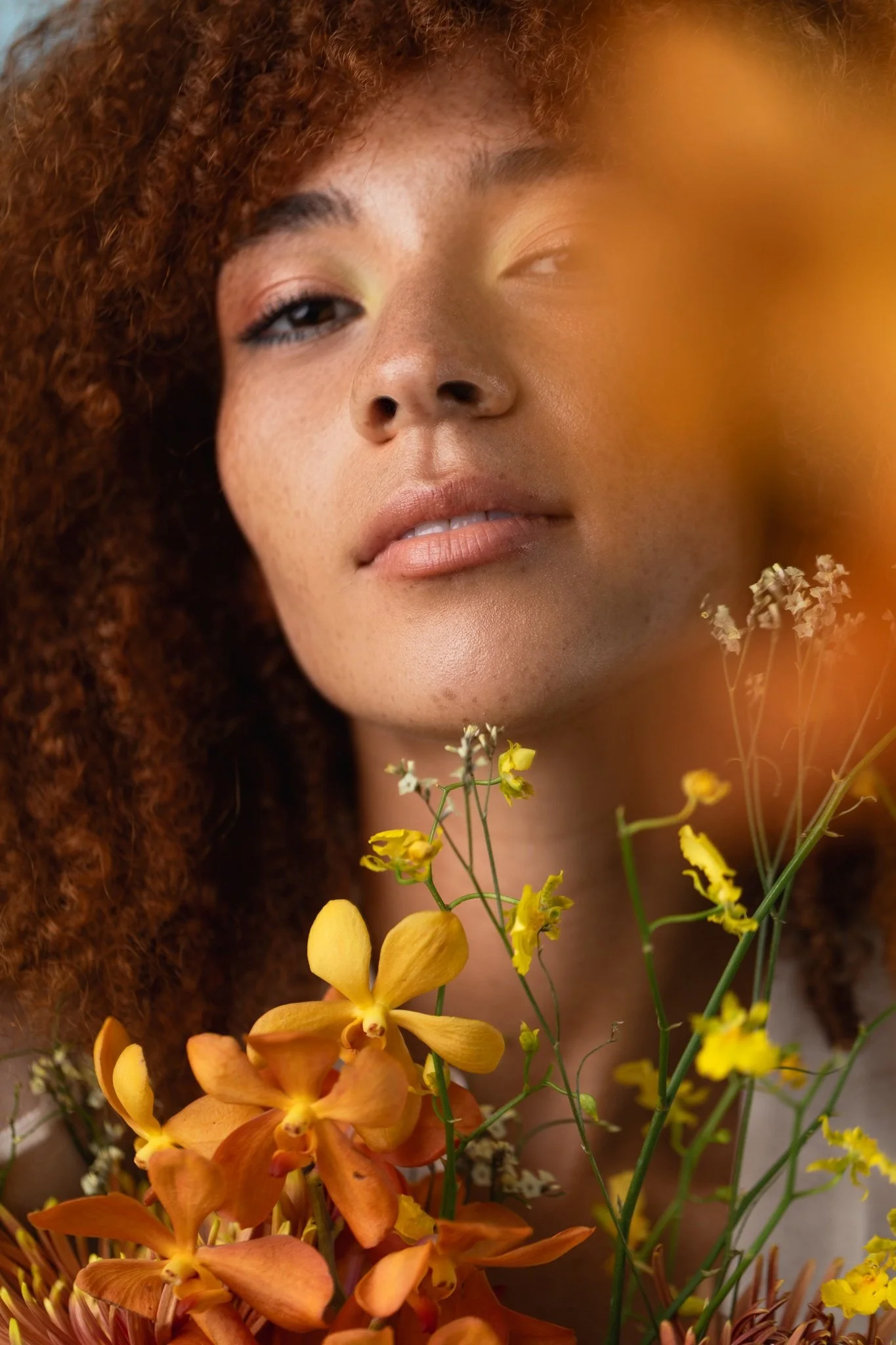 Close-up of a woman's face with curly red hair, surrounded by yellow and orange flowers.