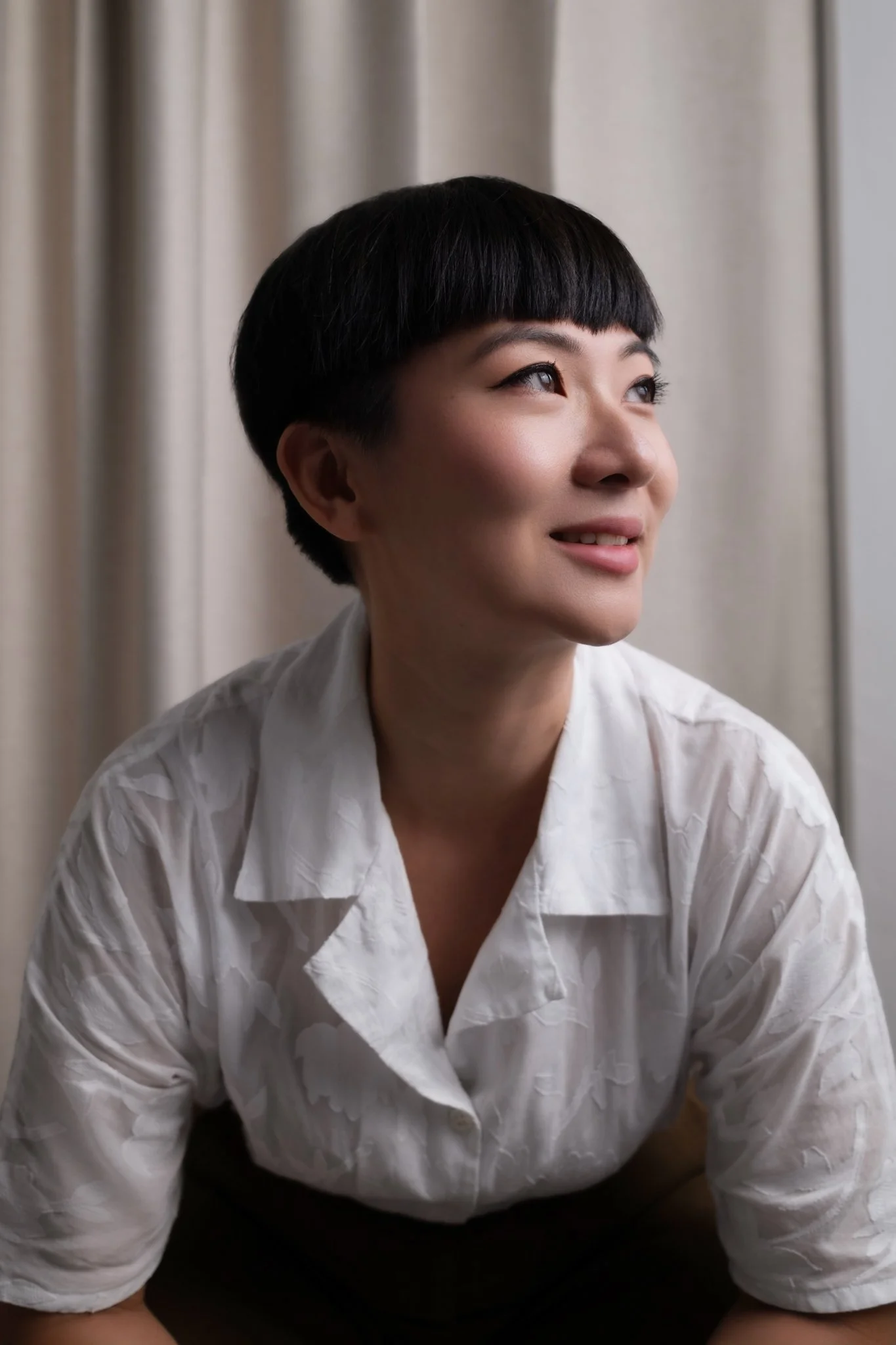 A woman with short black hair and a smile, wearing a white textured shirt, sitting indoors in front of beige curtains.