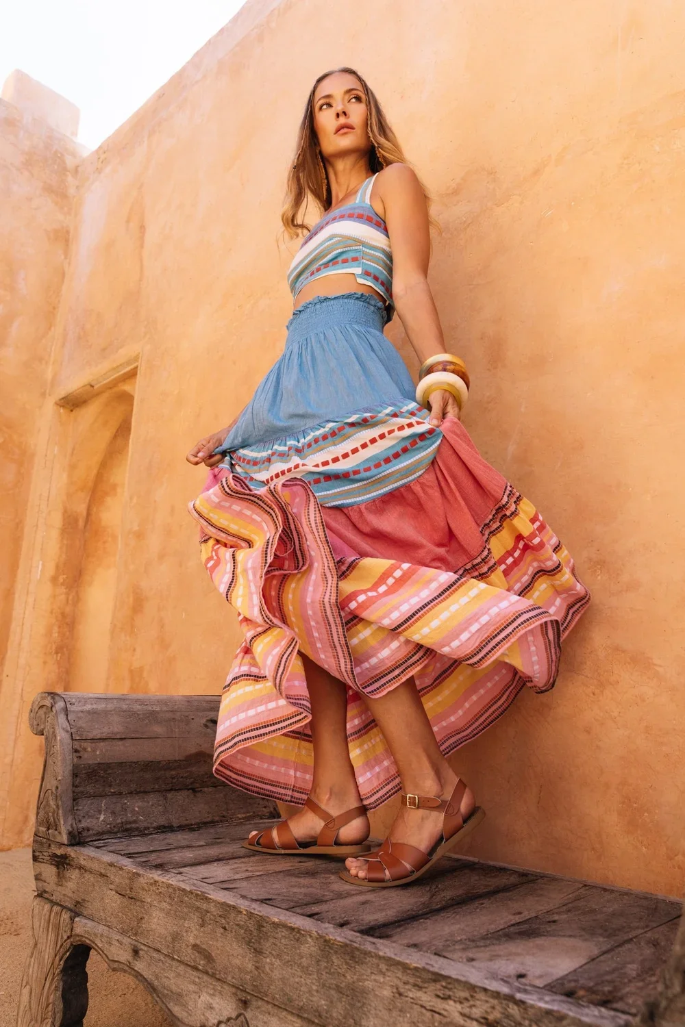 A woman in a colorful bohemian dress standing on a rustic wooden bench against a warm orange stucco wall.