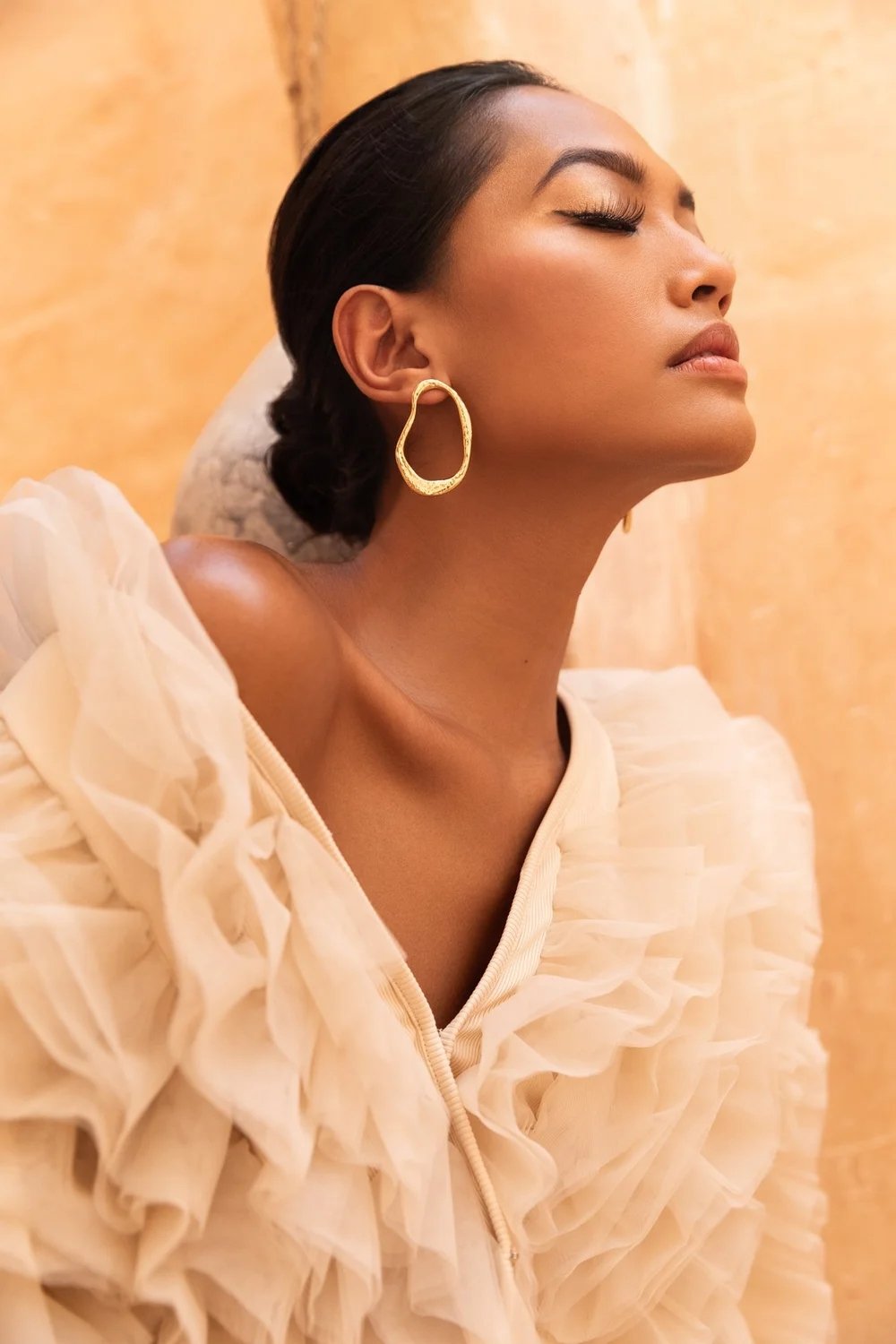 A woman with closed eyes and dark hair tied back, wearing a cream-colored ruffled dress and large gold earring, is resting against a beige background.