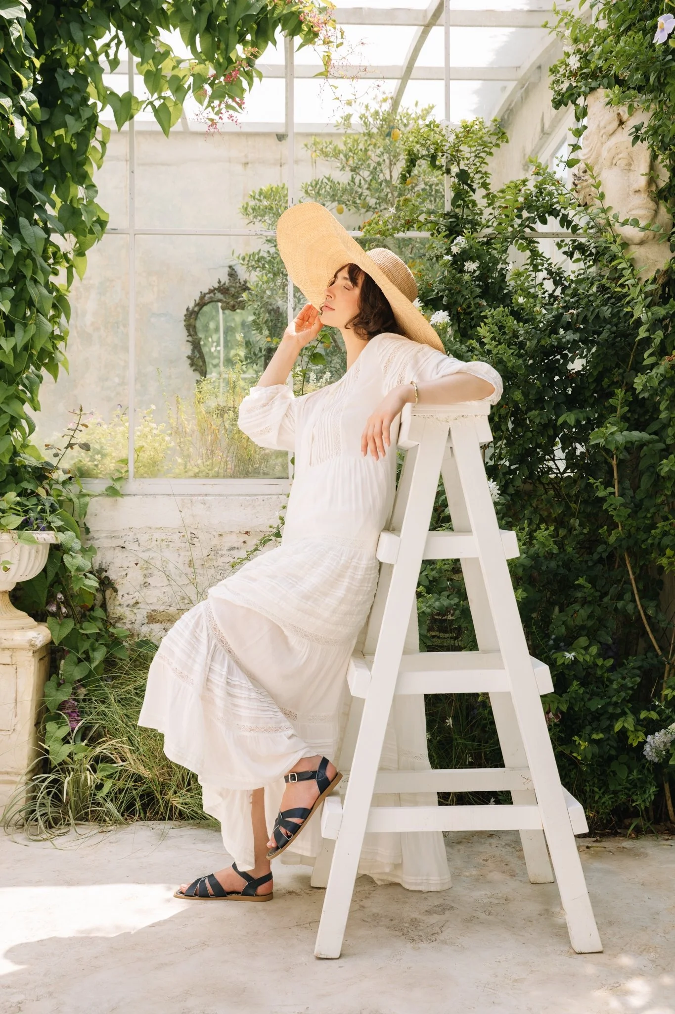 A woman sitting on a white step ladder in a greenhouse, wearing a white dress and large straw hat, with lush green plants and a window with a bust sculpture in the background.