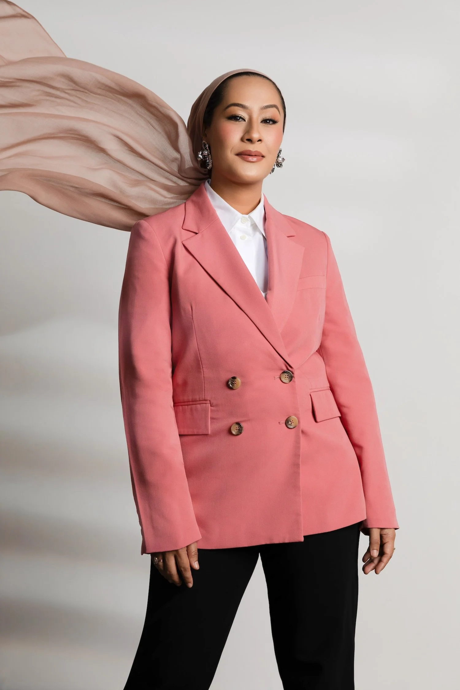 Woman wearing a pink blazer and black pants, with a light colored headscarf, standing against a plain background.