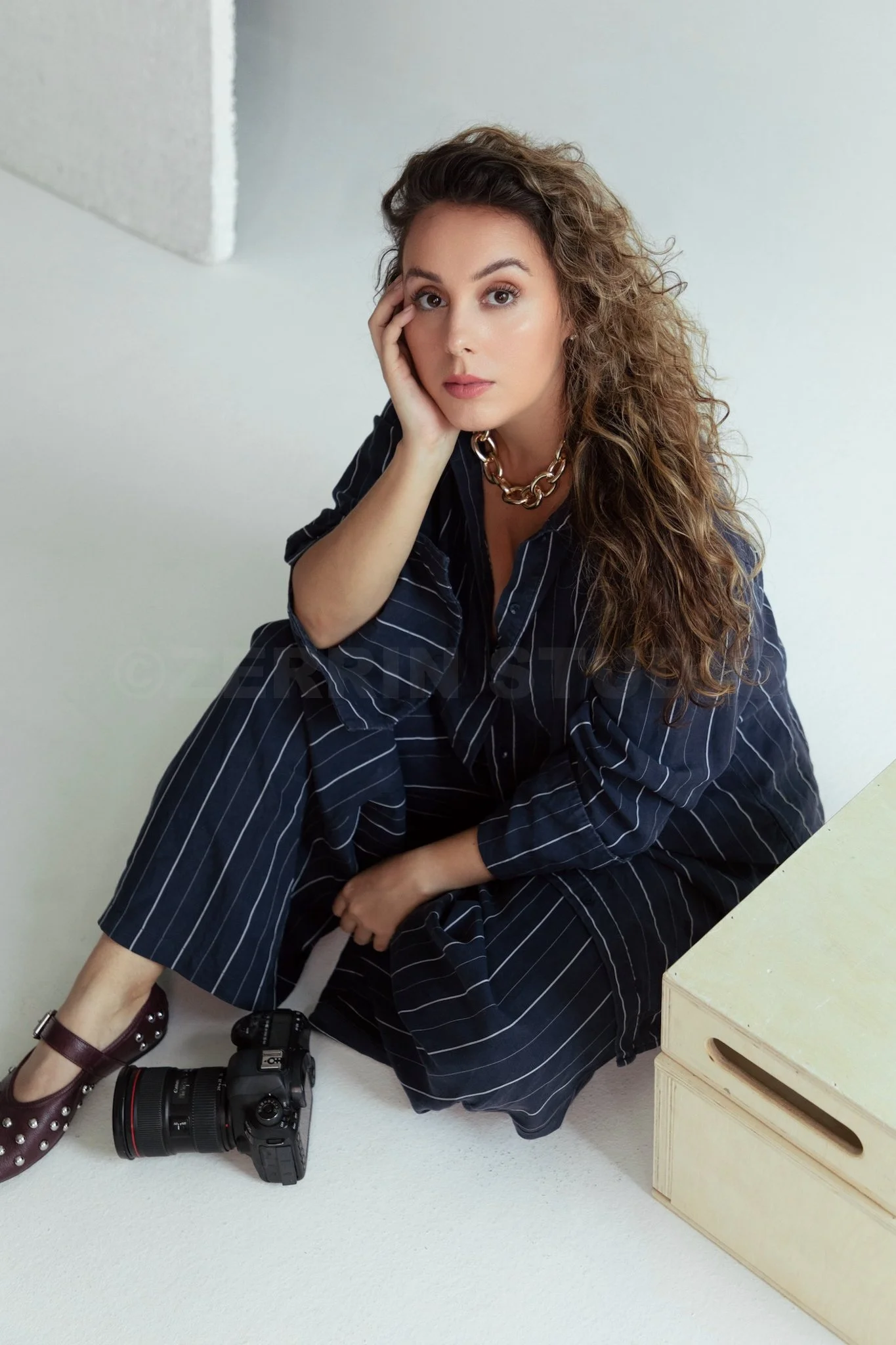 A woman with wavy brown hair, wearing a dark pinstripe dress, sitting on the floor with a camera beside her and a wooden box nearby.