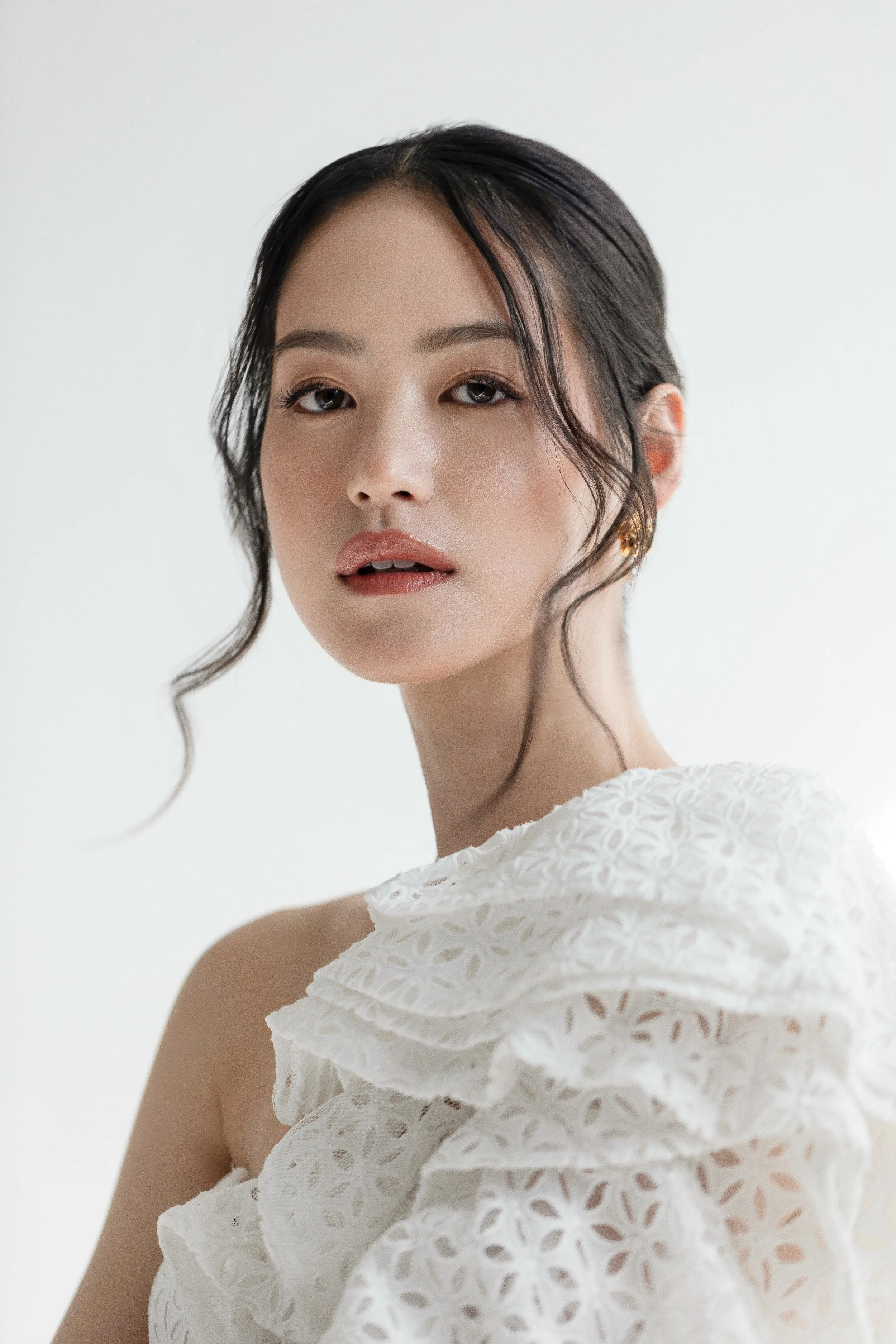 A young woman with dark hair styled in loose waves, wearing subtle makeup and a white lace dress with ruffles, looking at the camera with a soft expression against a plain light background.