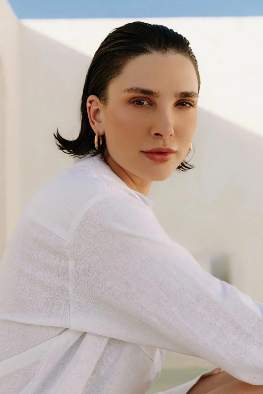 A woman with short, dark, wet-looking hair, wearing gold hoop earrings and a white top, sitting outdoors next to a white wall with a blue sky background.