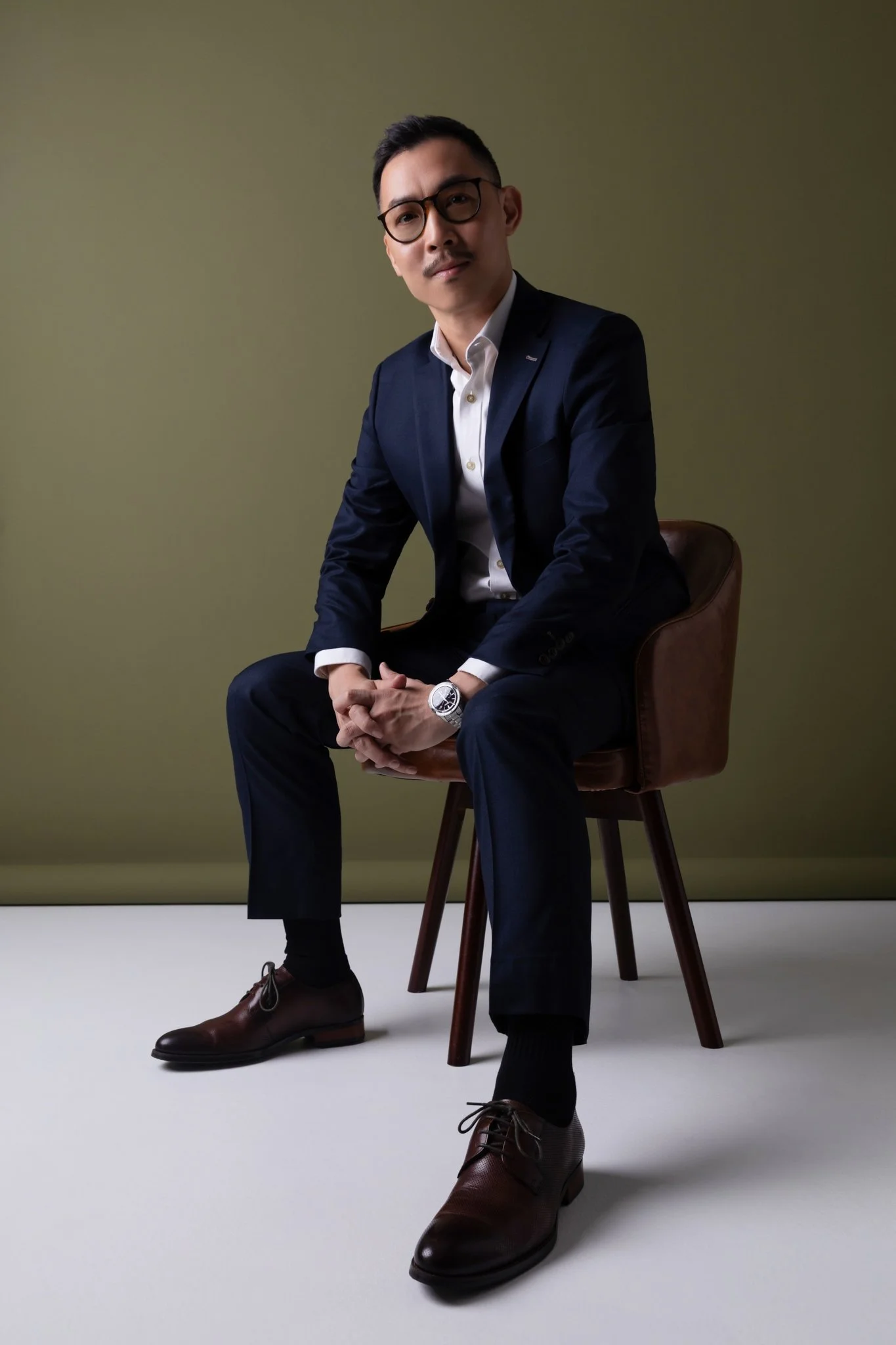 A man wearing glasses, a dark suit, and a white shirt seated on a wooden chair against a plain background, looking at the camera.
