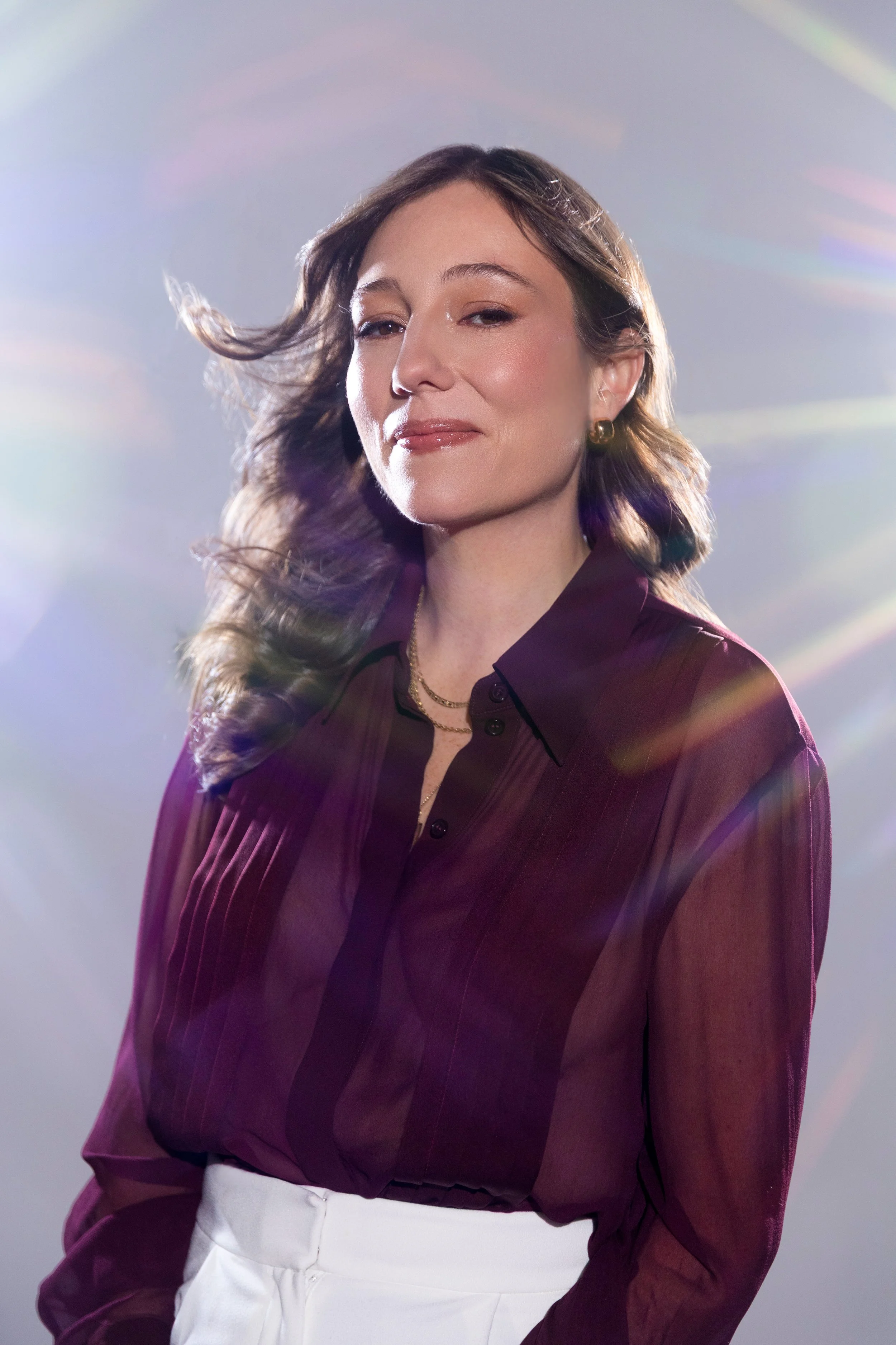 A woman with wavy brown hair, wearing a maroon blouse and white pants, standing against a light grey background with rainbow lens flare effects.
