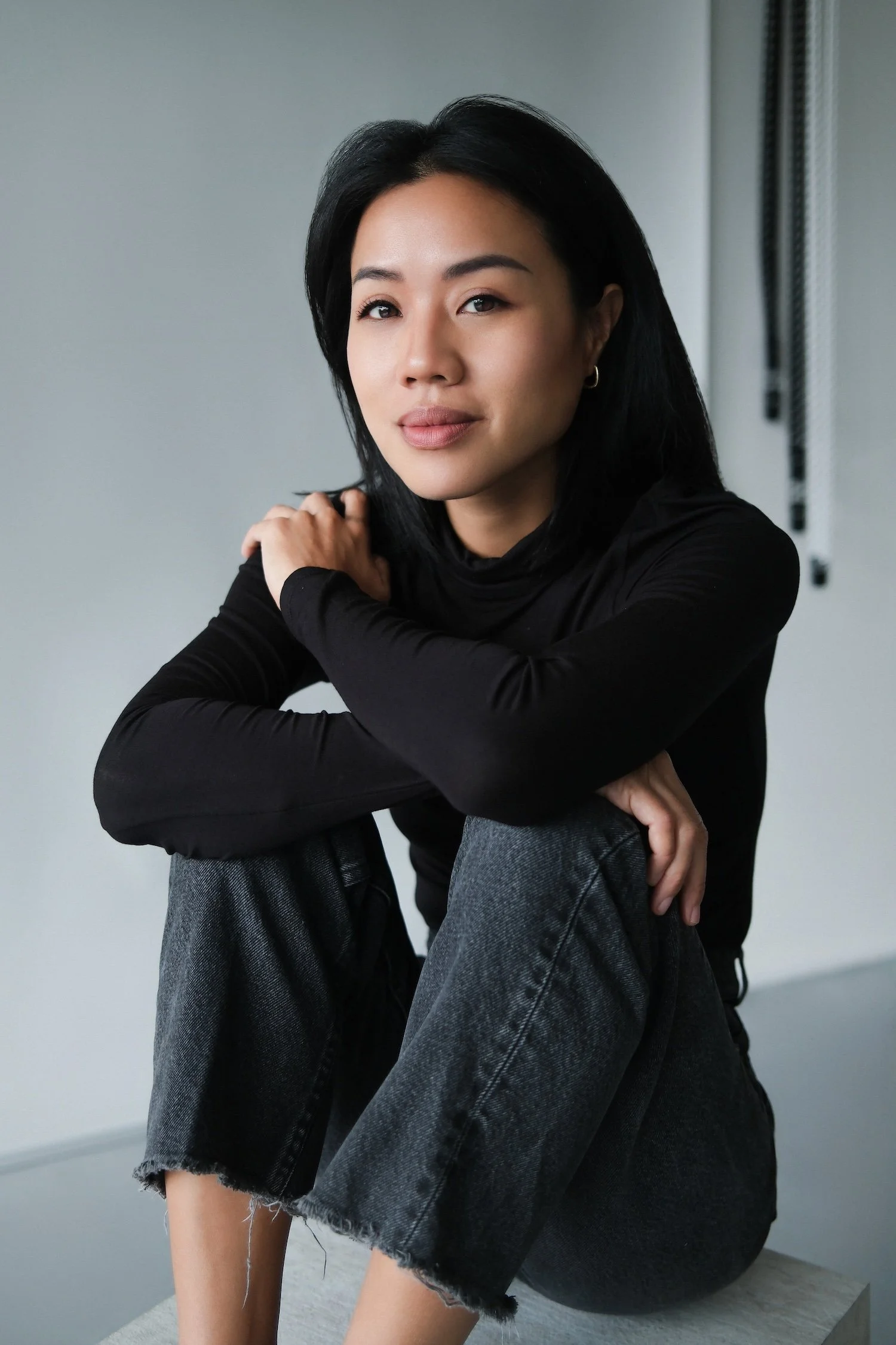 A woman with short black hair, wearing a black long-sleeve shirt and gray jeans, sitting on a gray surface in front of a plain light-colored wall, with her arms crossed on her knee, looking at the camera.
