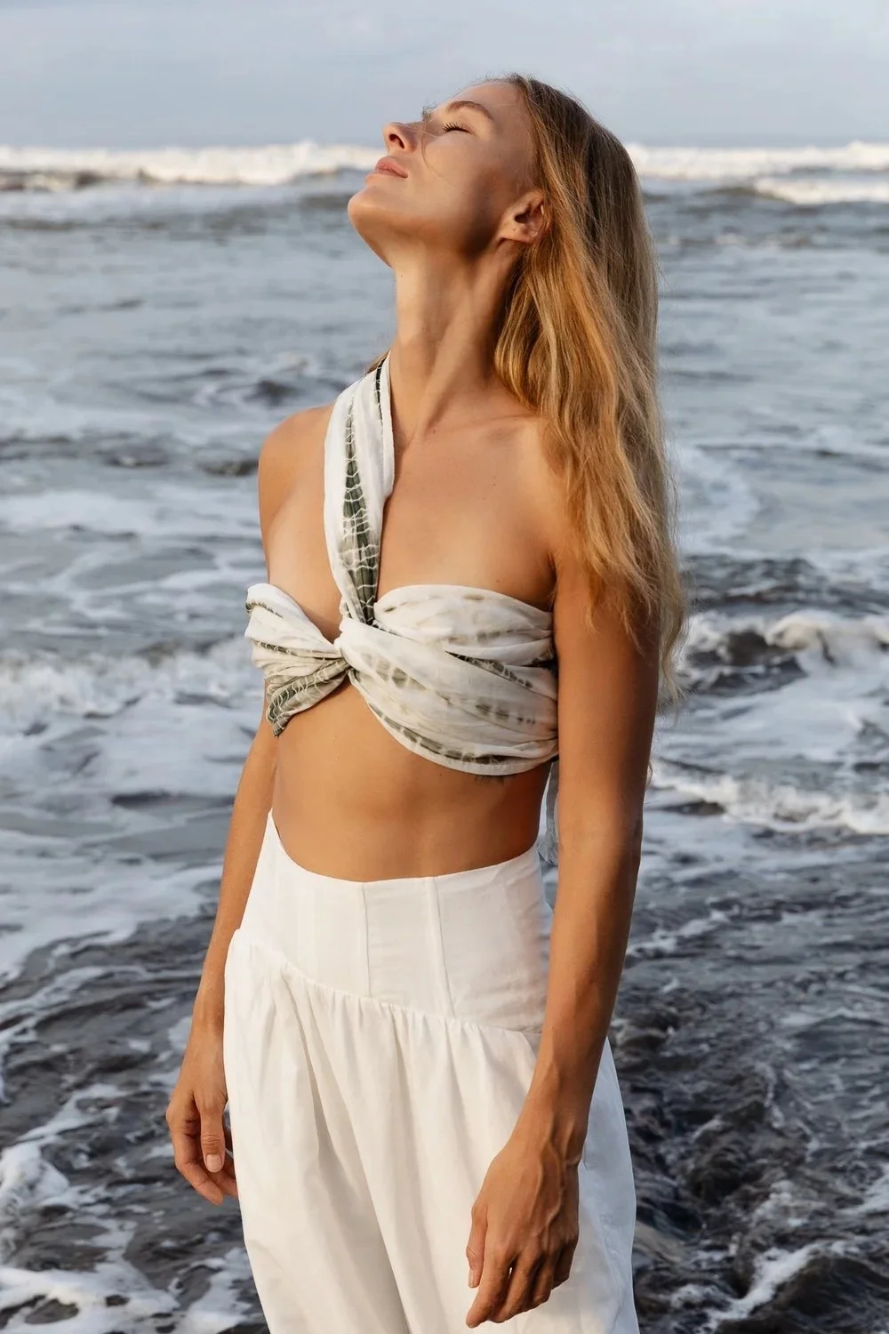 Woman standing on the beach with eyes closed and face tilted upward, wearing a white and green tie-dye wrap top and white high-waisted skirt, ocean waves in the background.