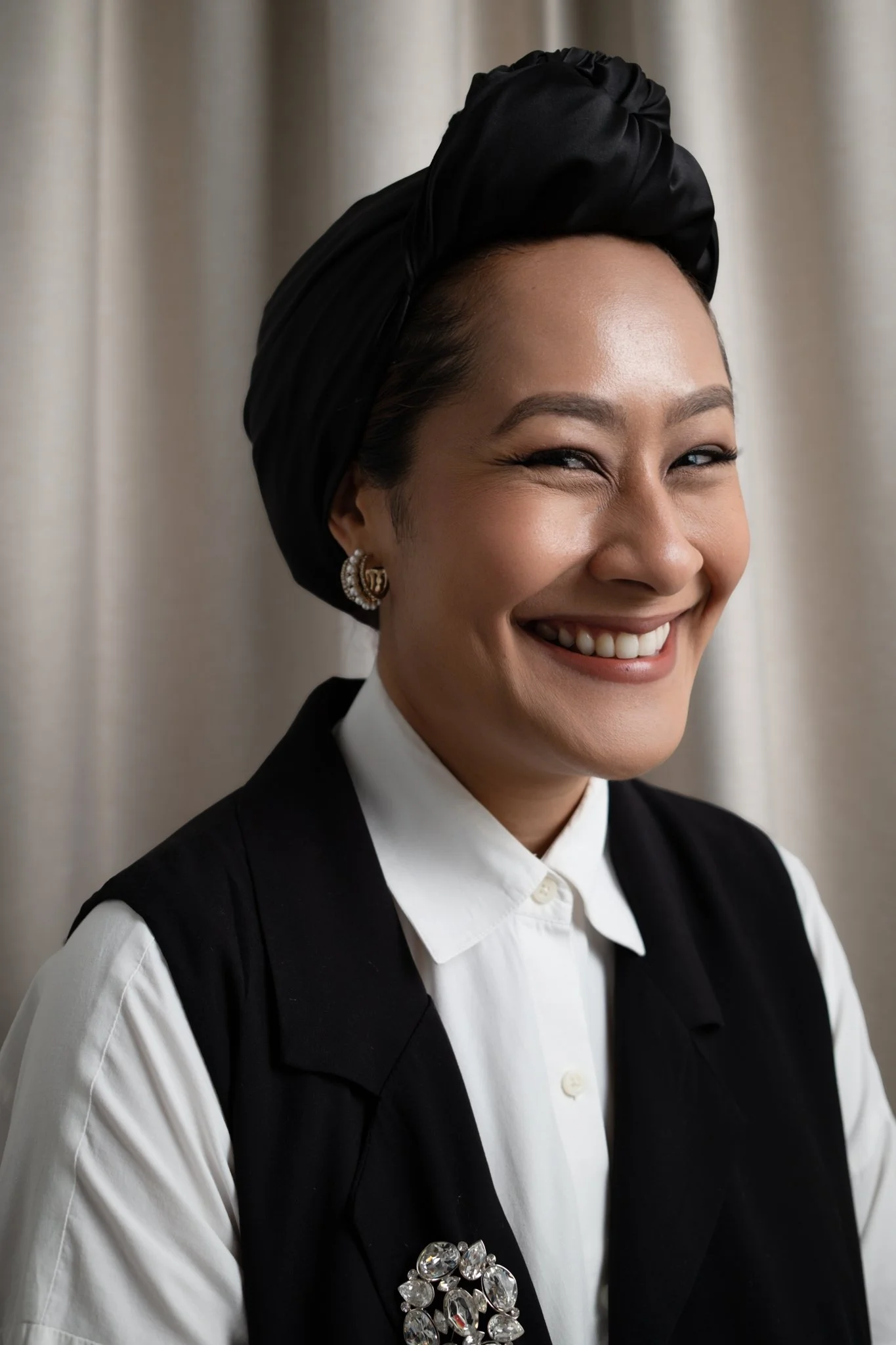 A woman with a bright smile, wearing a black headwrap, earrings, a white shirt, and a black vest with some decorative jewels on the left side, standing in front of a neutral curtain.