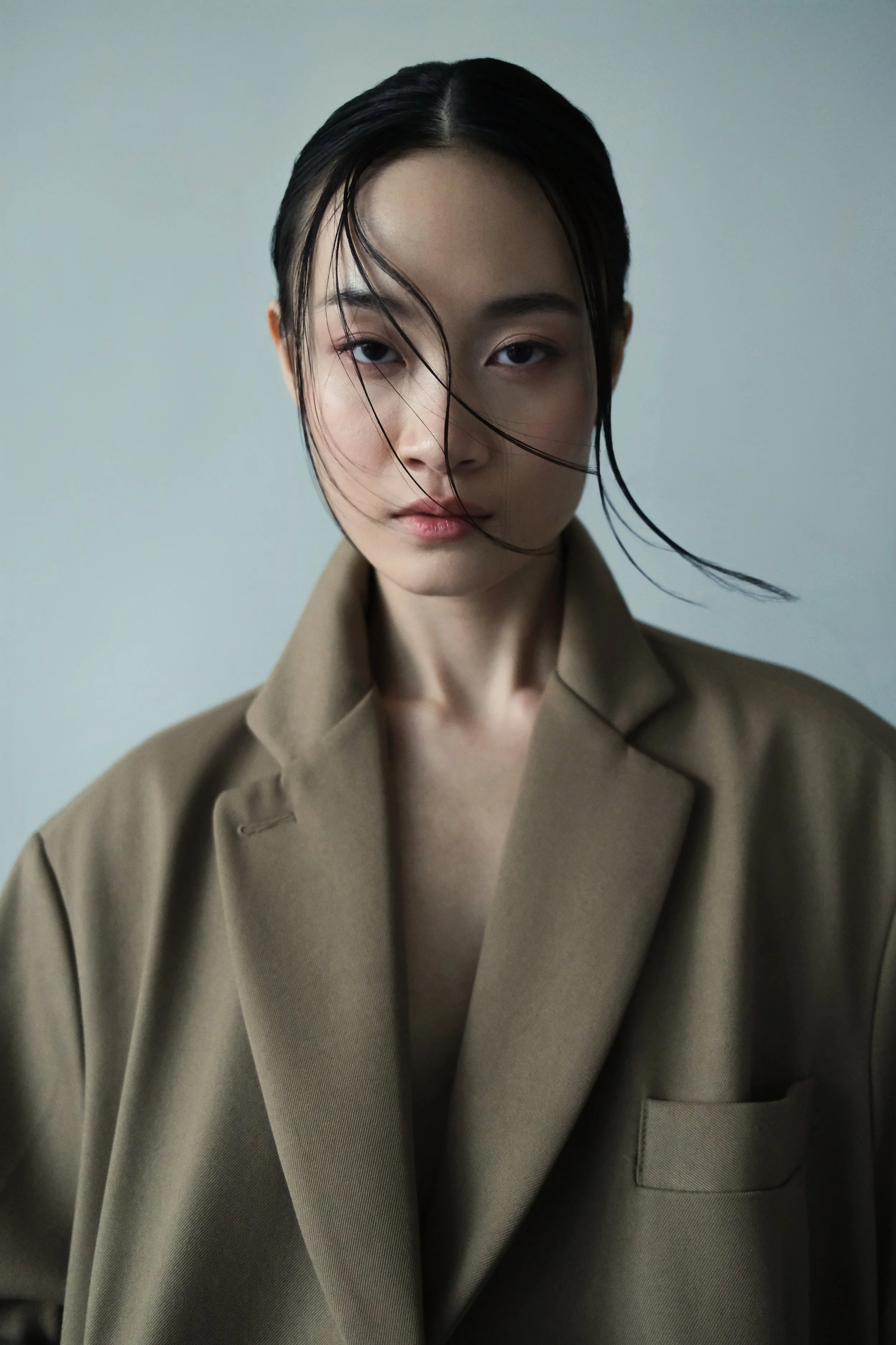 A portrait of a woman with dark hair, some of which is wet and falling across her face, dressed in a beige trench coat, against a plain light gray background.
