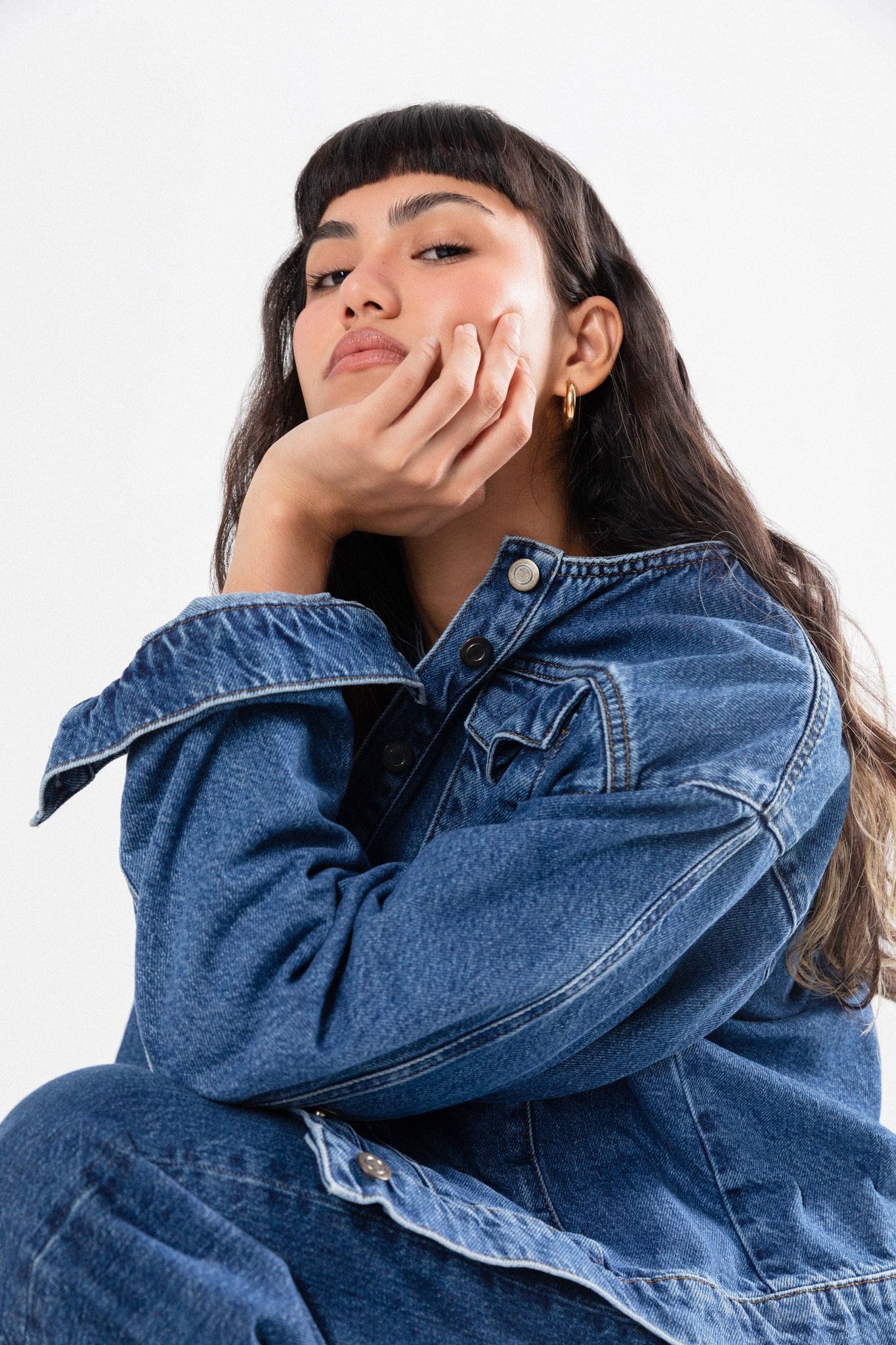 A young woman with dark hair, wearing a denim jacket and gold hoop earrings, resting her chin on her hand and looking confidently at the camera against a plain white background.