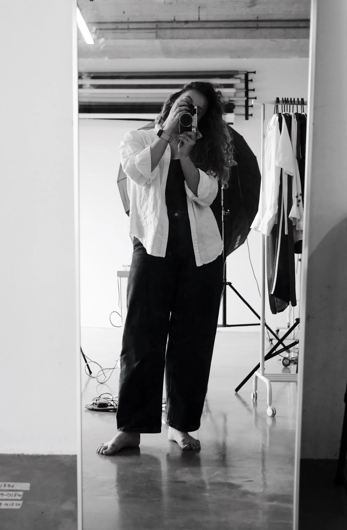 A woman with long, curly hair taking a selfie in a large mirror. She is standing barefoot on a wooden floor, wearing a light-colored top over a darker shirt and dark pants. The background shows photography equipment, including a lighting umbrella, clothing rack, and various lighting or filming stands.