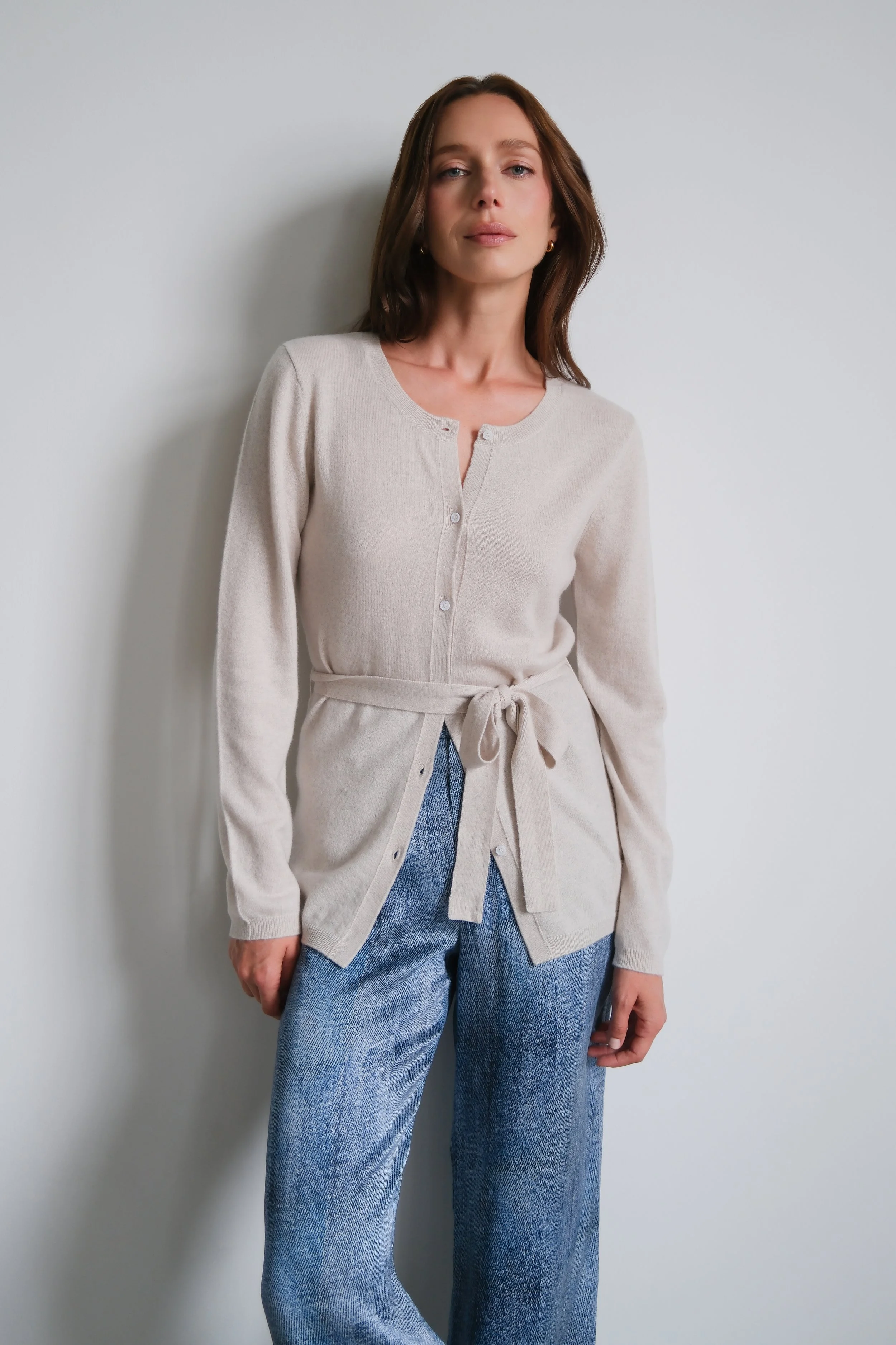A woman with brown hair wearing a beige cardigan tied at the waist and blue jeans, standing against a light grey wall.