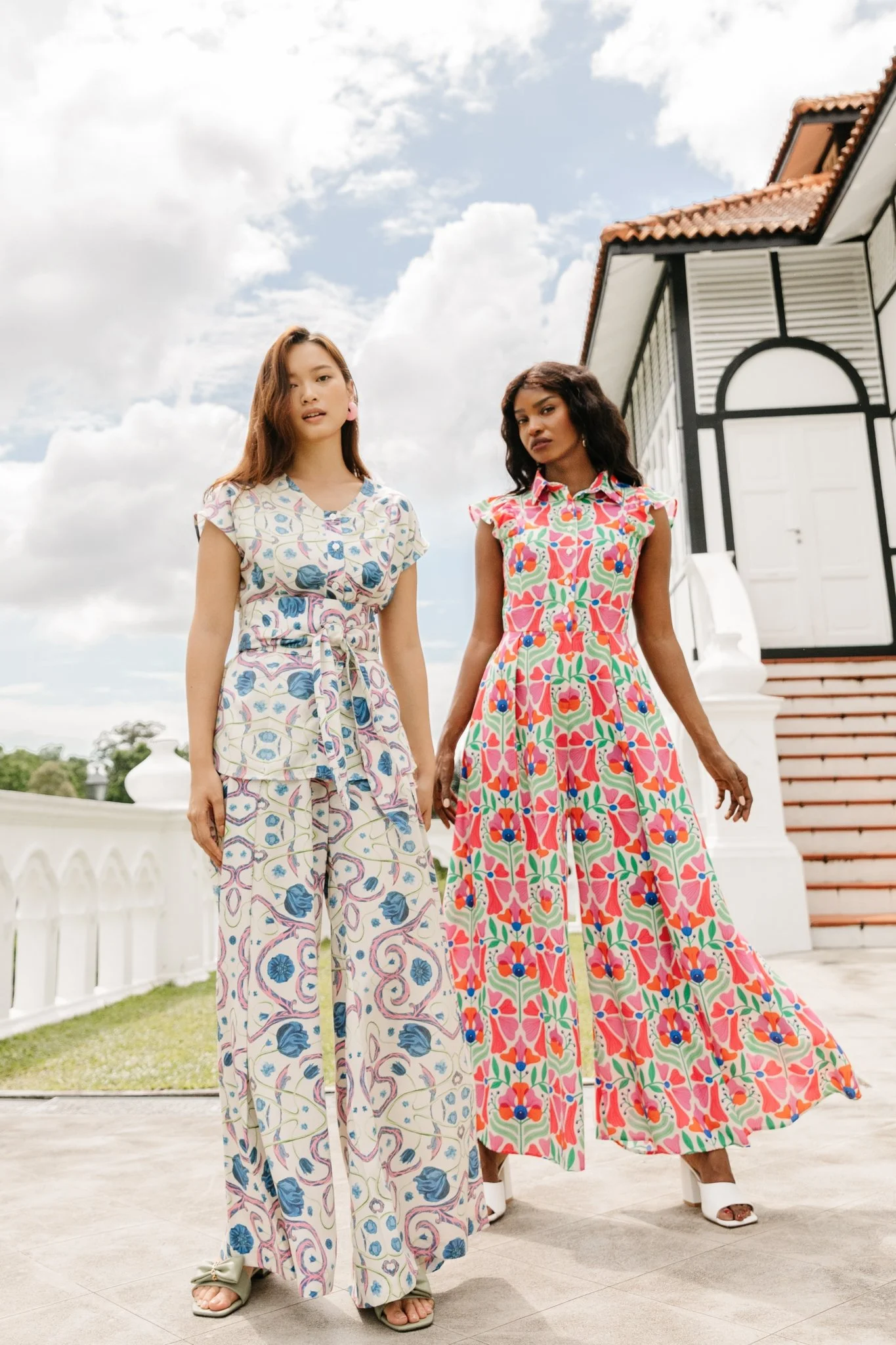 Two women standing in front of a white building with a red tiled roof, wearing colorful patterned maxi dresses, under a partly cloudy sky.