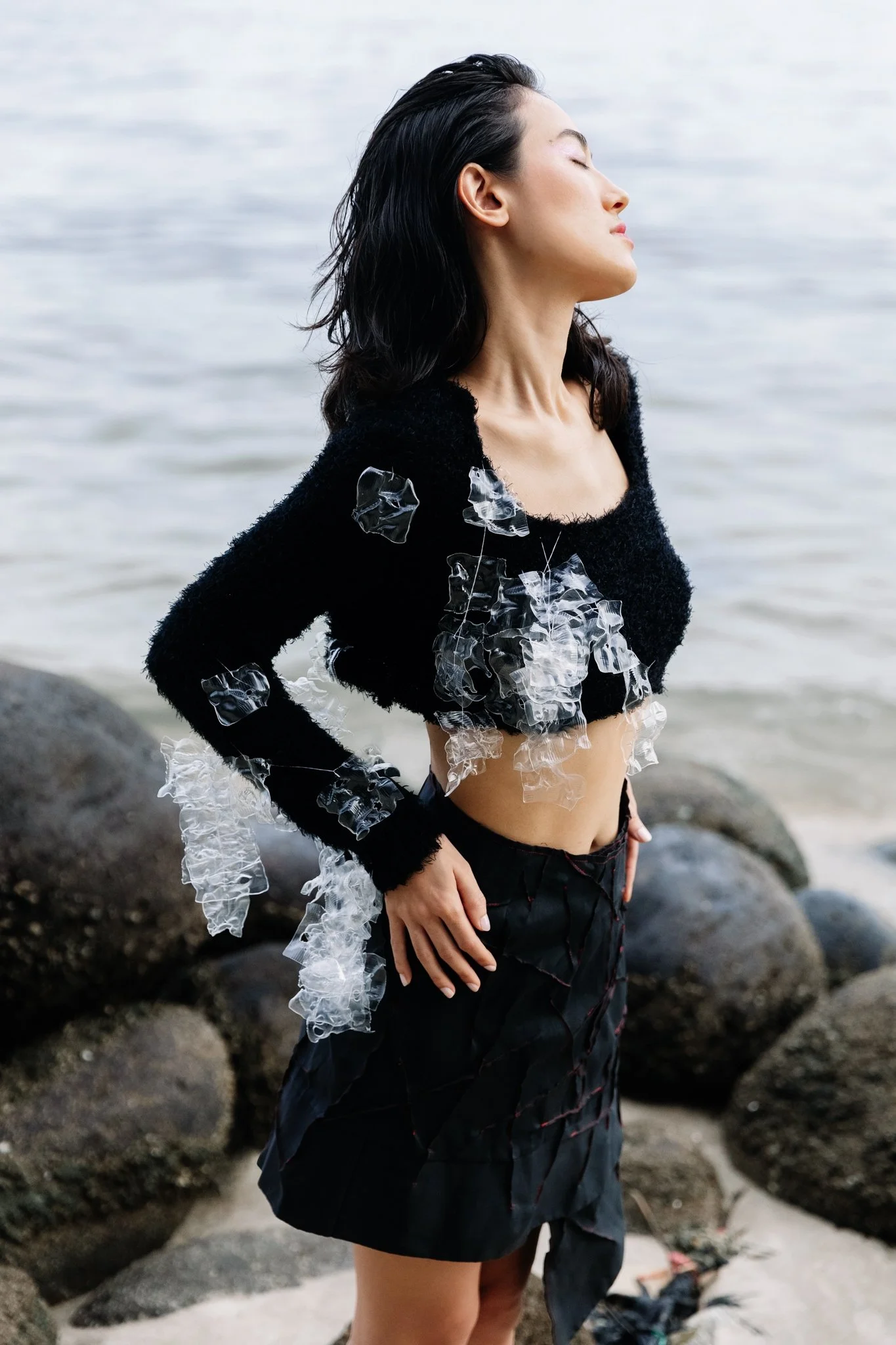 A woman with long dark hair and light skin stands by the water on a rocky beach, wearing a black fuzzy top with transparent plastic decorative accents, and a black skirt with red stitching.