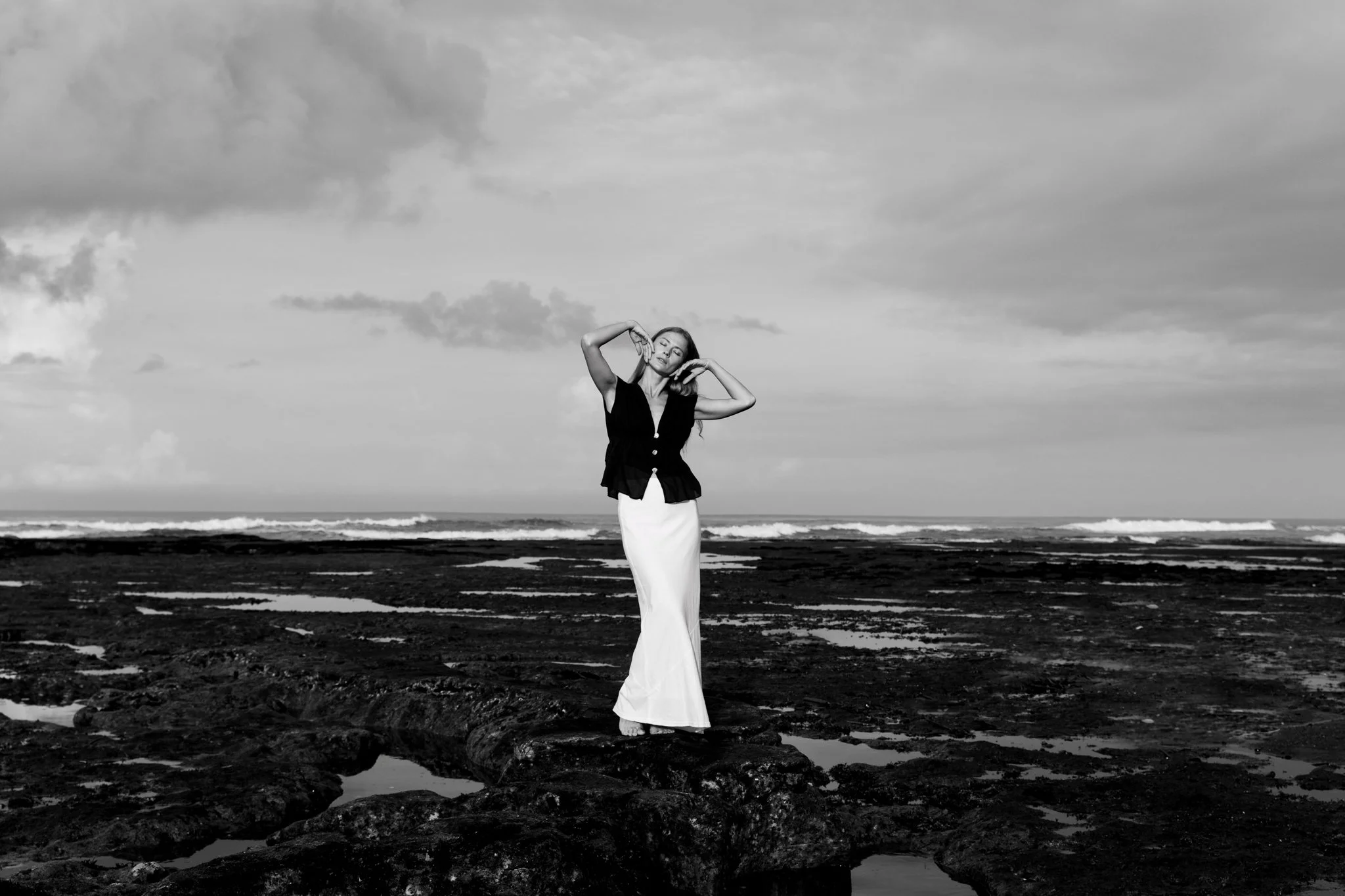 A woman in a black top and white skirt standing on rocks at the edge of the ocean with a cloudy sky in black and white.