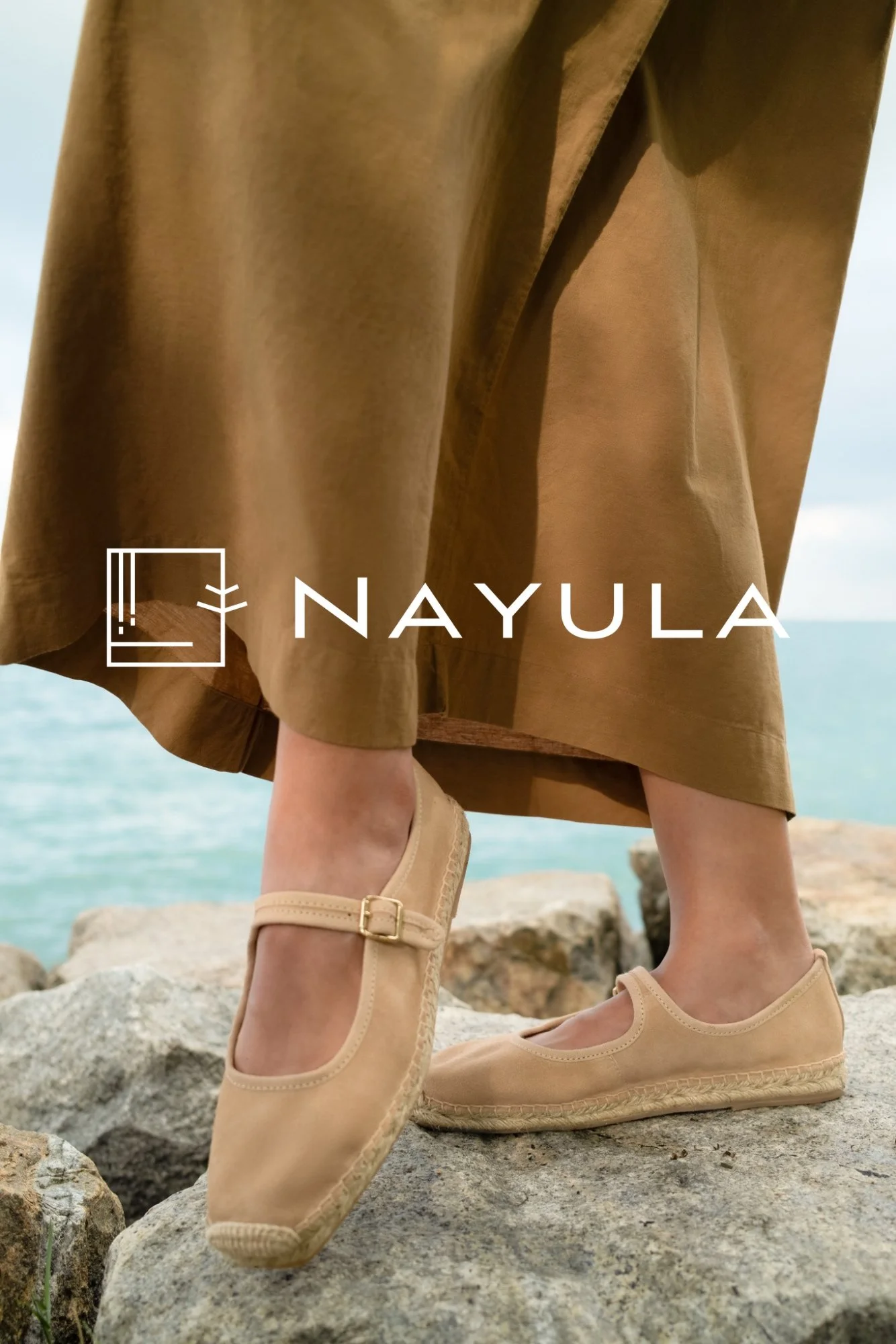 Close-up of a person standing on rocks near the water, wearing beige shoes, a brown skirt, and a brown top. The background shows a blurred view of the sea and sky.