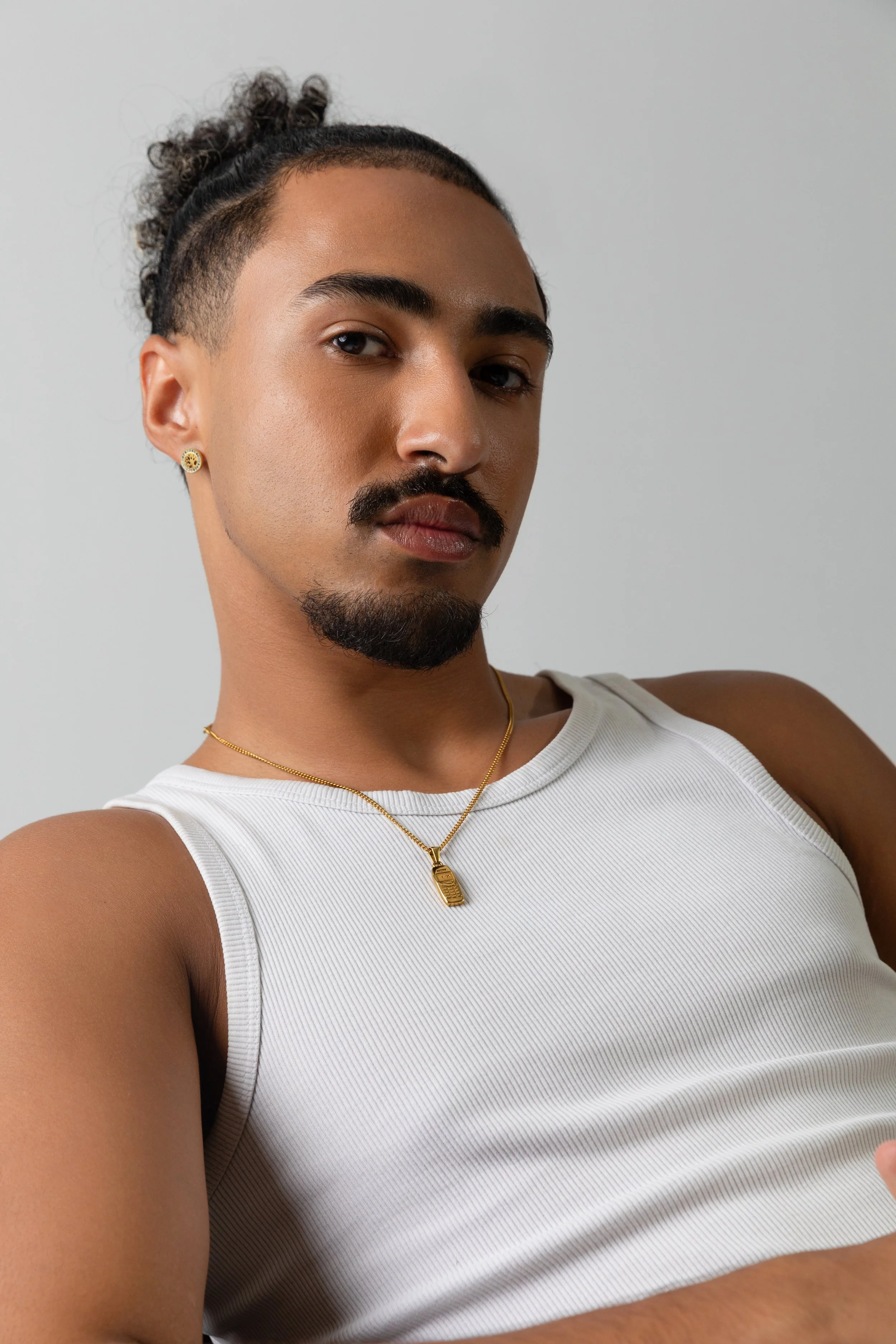 Close-up portrait of a person with well-groomed facial hair, gold jewelry, and a white sleeveless shirt, against a plain gray background.