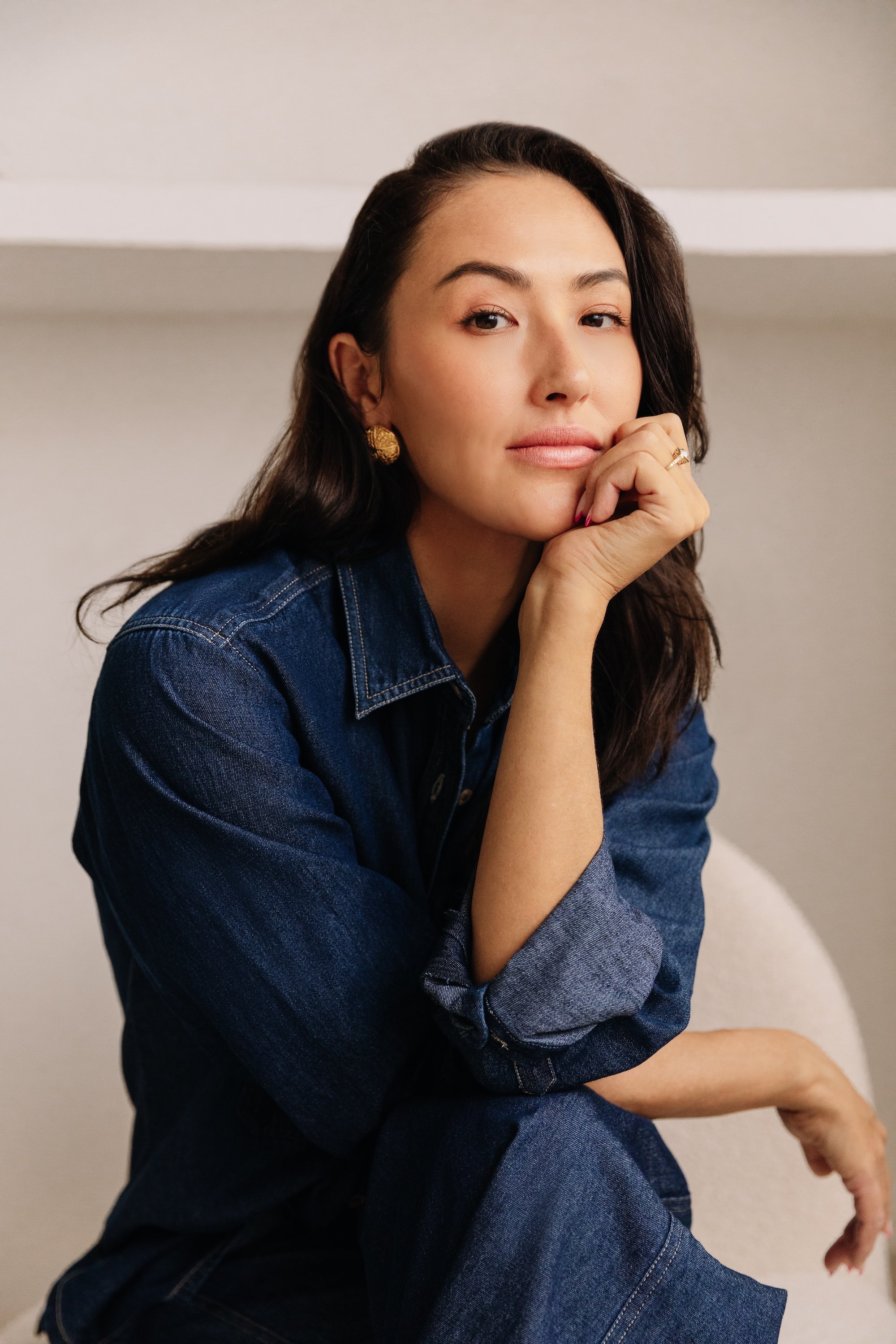 A woman with long dark hair, wearing a denim jacket and gold earrings, resting her chin on her hand and sitting on a chair with a neutral background.