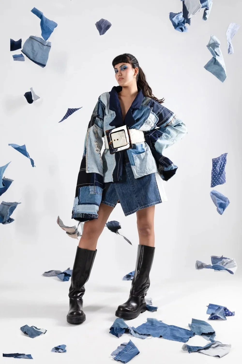 Fashion model stands confidently surrounded by floating torn pieces of denim and fabric, wearing a patchwork denim jacket, matching skirt, and tall black boots.