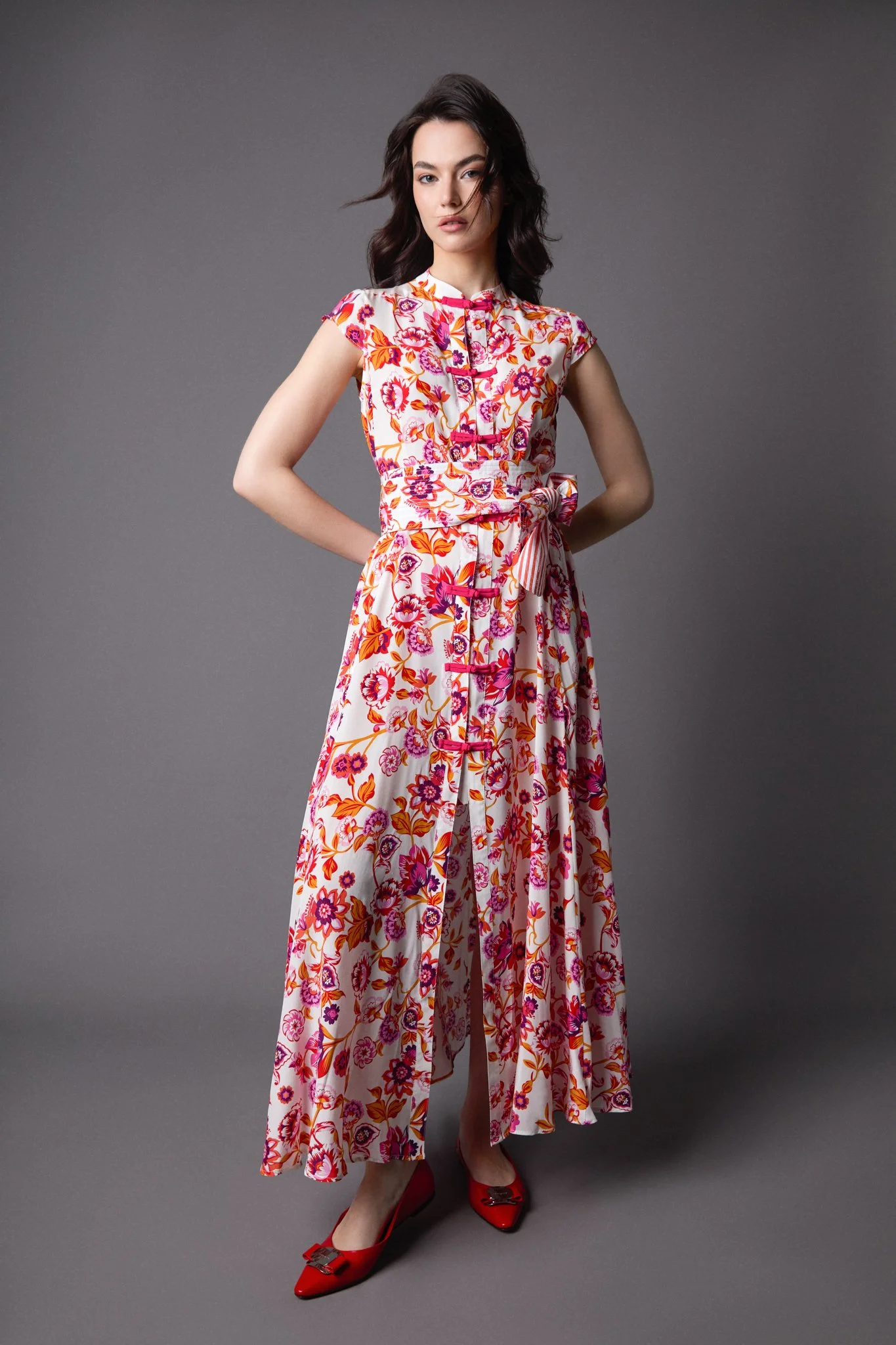 A woman dressed in a floral dress with a white background, standing against a plain gray backdrop, wearing red high heels.