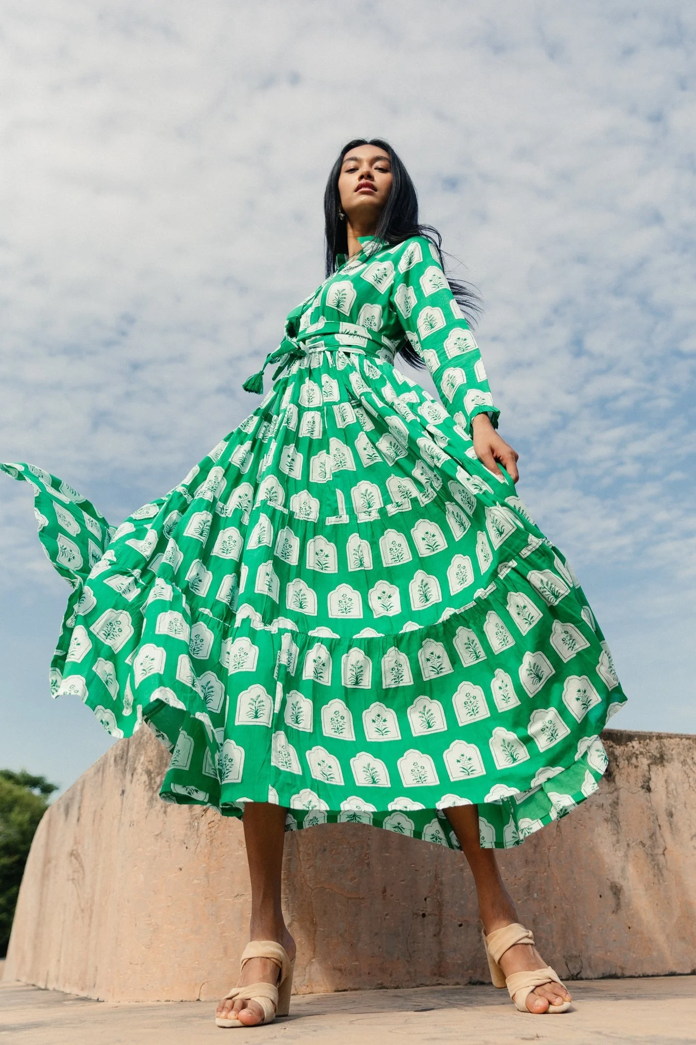 A woman in a long green patterned dress with white designs is standing outdoors on a stone surface, with her legs apart and hands adjusting her dress. She is wearing beige high-heeled sandals and has long dark hair. The sky in the background is cloud