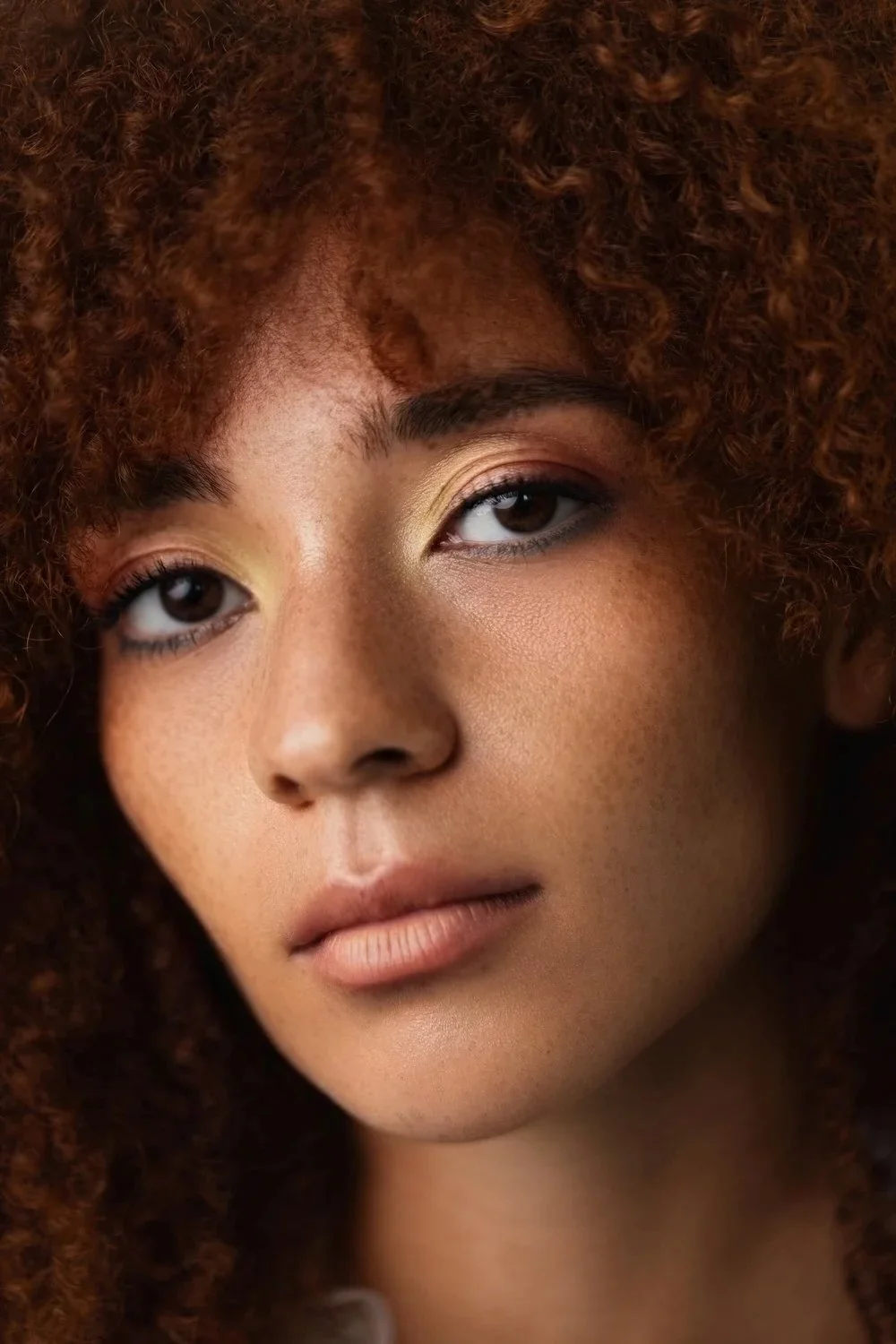 Close-up of a woman with curly red hair, subtle makeup, and neutral expression.