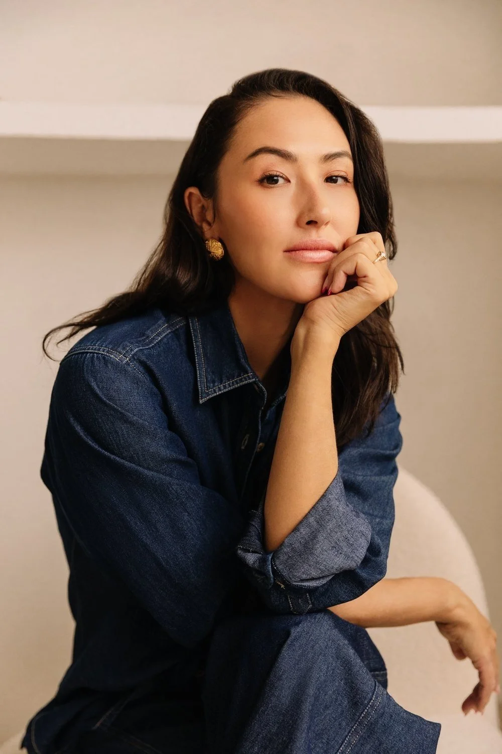 A woman with dark hair, wearing a denim jacket, sitting and resting her chin on her hand, looking at the camera.
