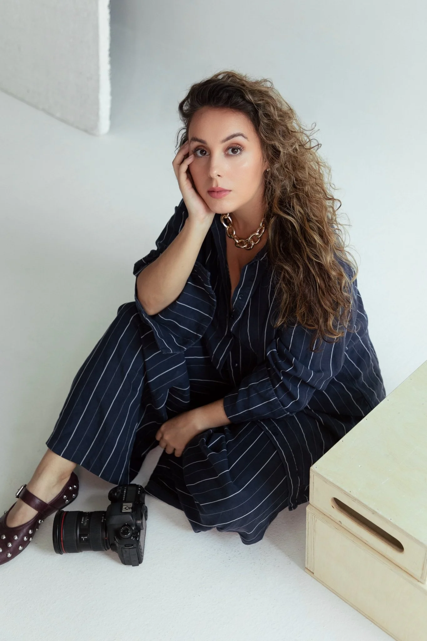 A woman with curly brown hair, sitting on the floor next to a camera, wearing a dark striped shirt and jewelry.