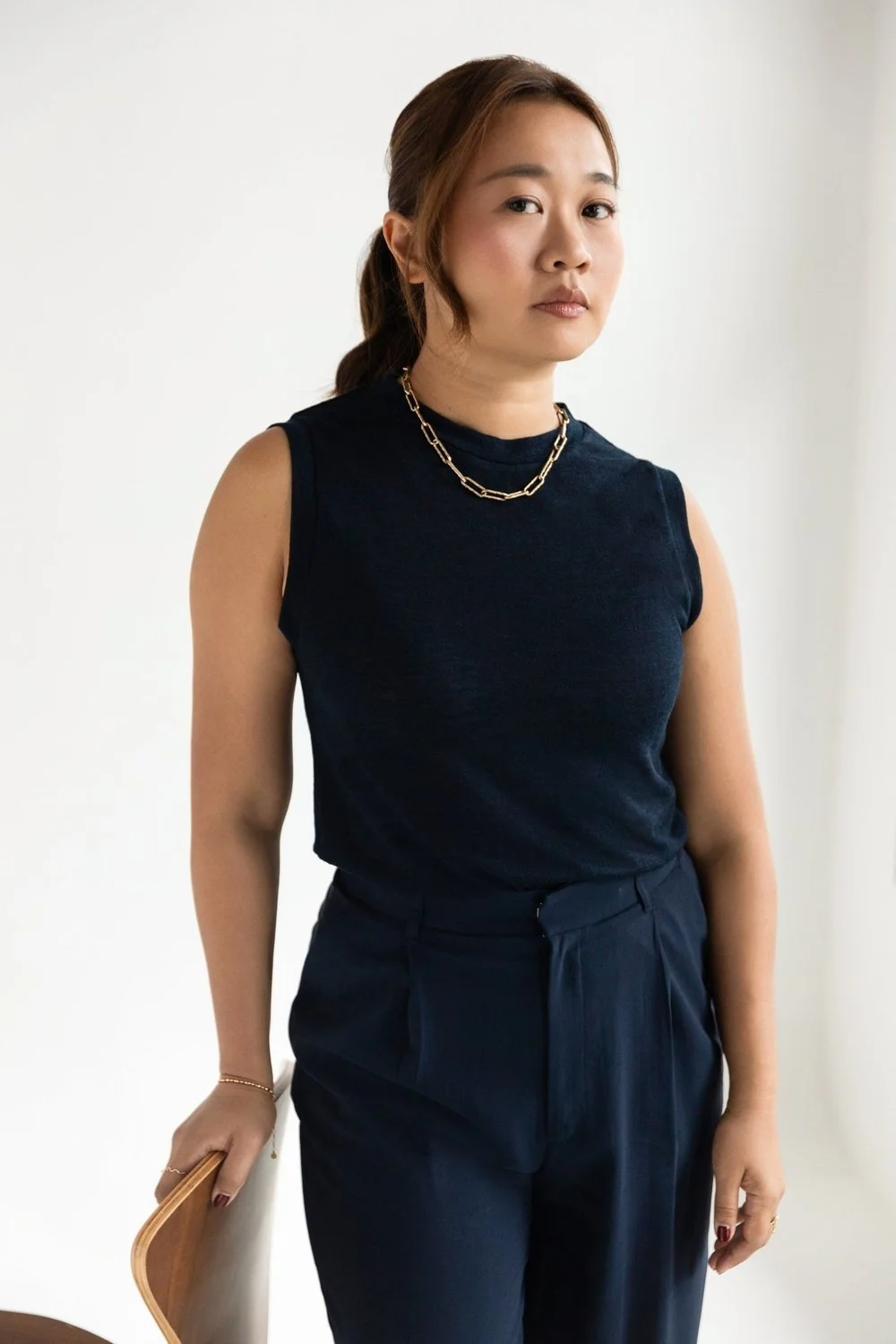 A woman with brown hair in a ponytail, wearing a sleeveless black top, black pants, and a gold chain necklace, standing indoors against a white wall.