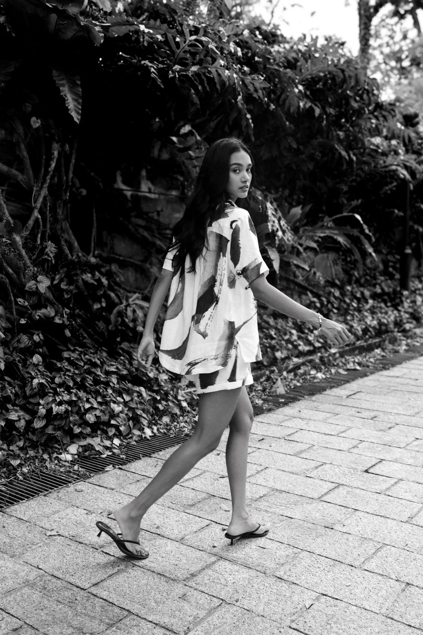 A woman with long dark hair walking along a stone path next to a hedge, wearing a loose dress with a bold abstract pattern and flip-flops.