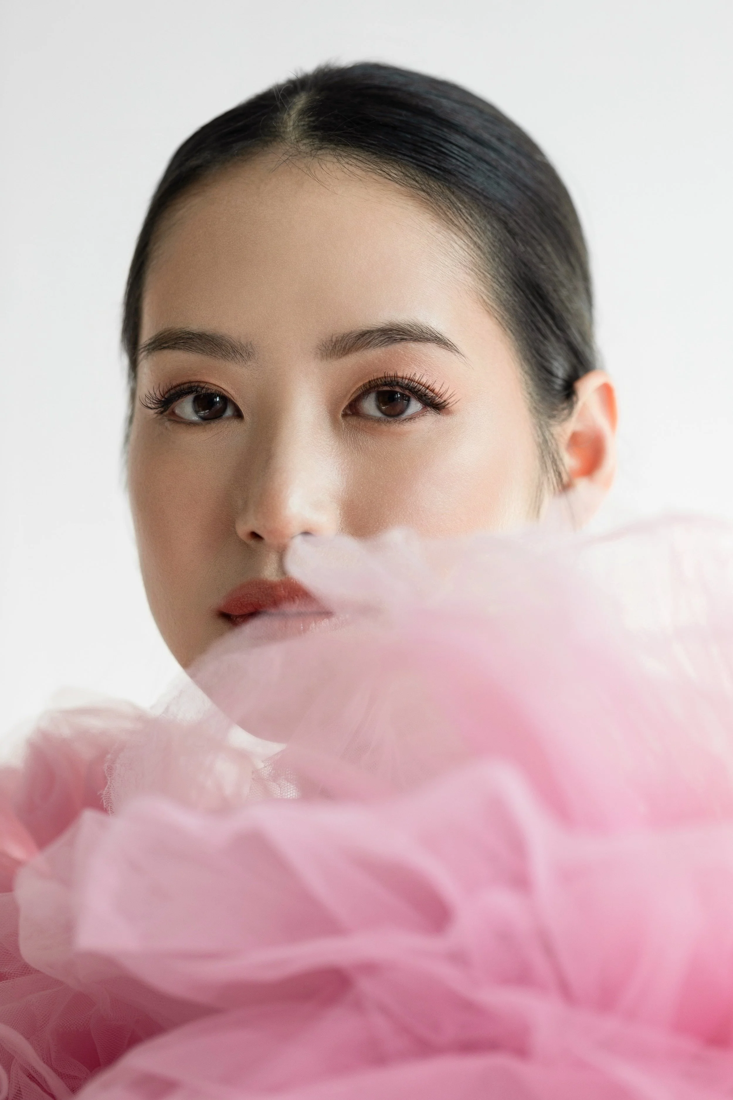 Close-up of a woman's face with makeup, partially obscured by pink peony flowers in the foreground, soft lighting, white background.