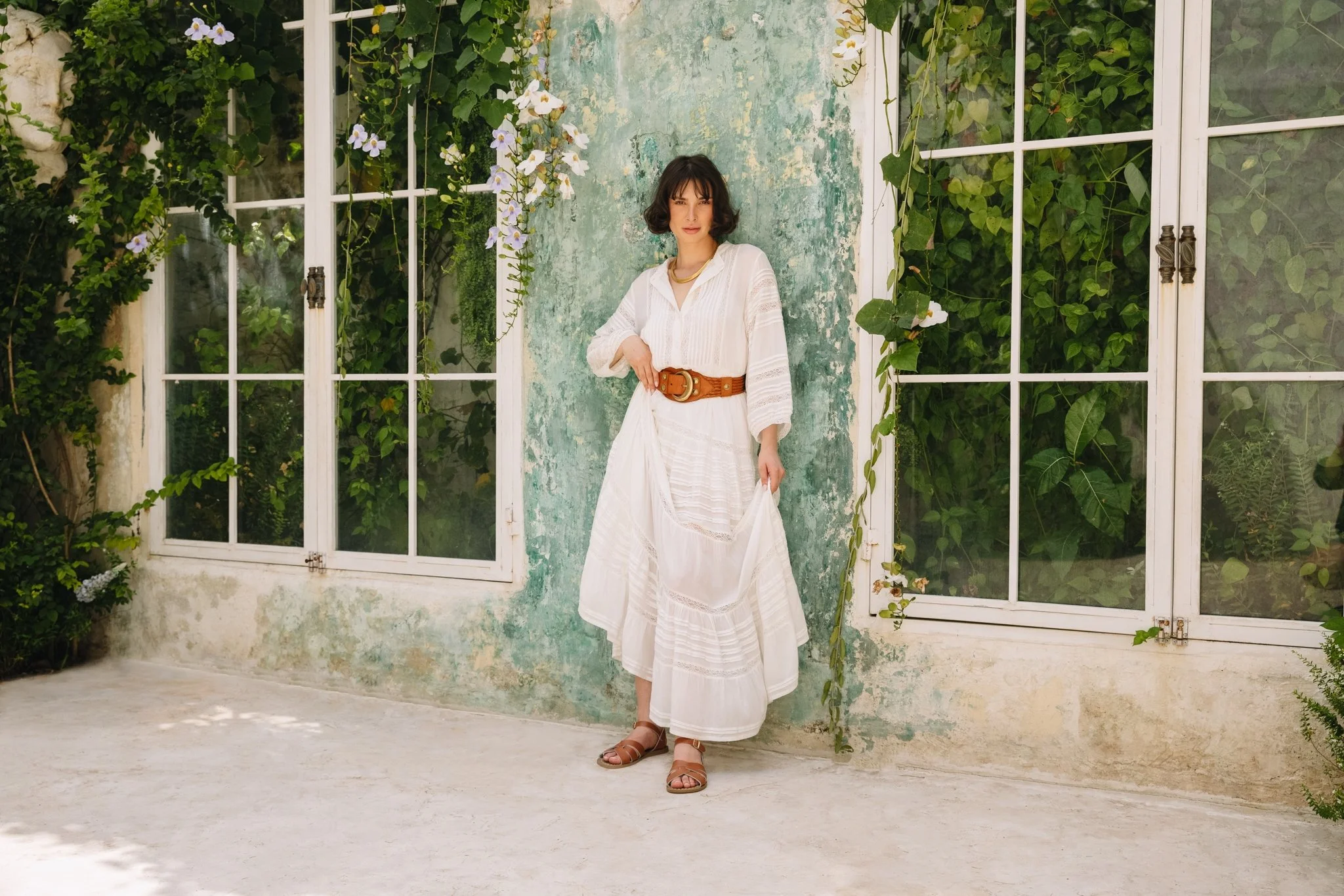 A woman in a white bohemian-style dress and brown sandals standing in front of a rustic wall with large glass windows and green foliage with climbing plants and white flowers.