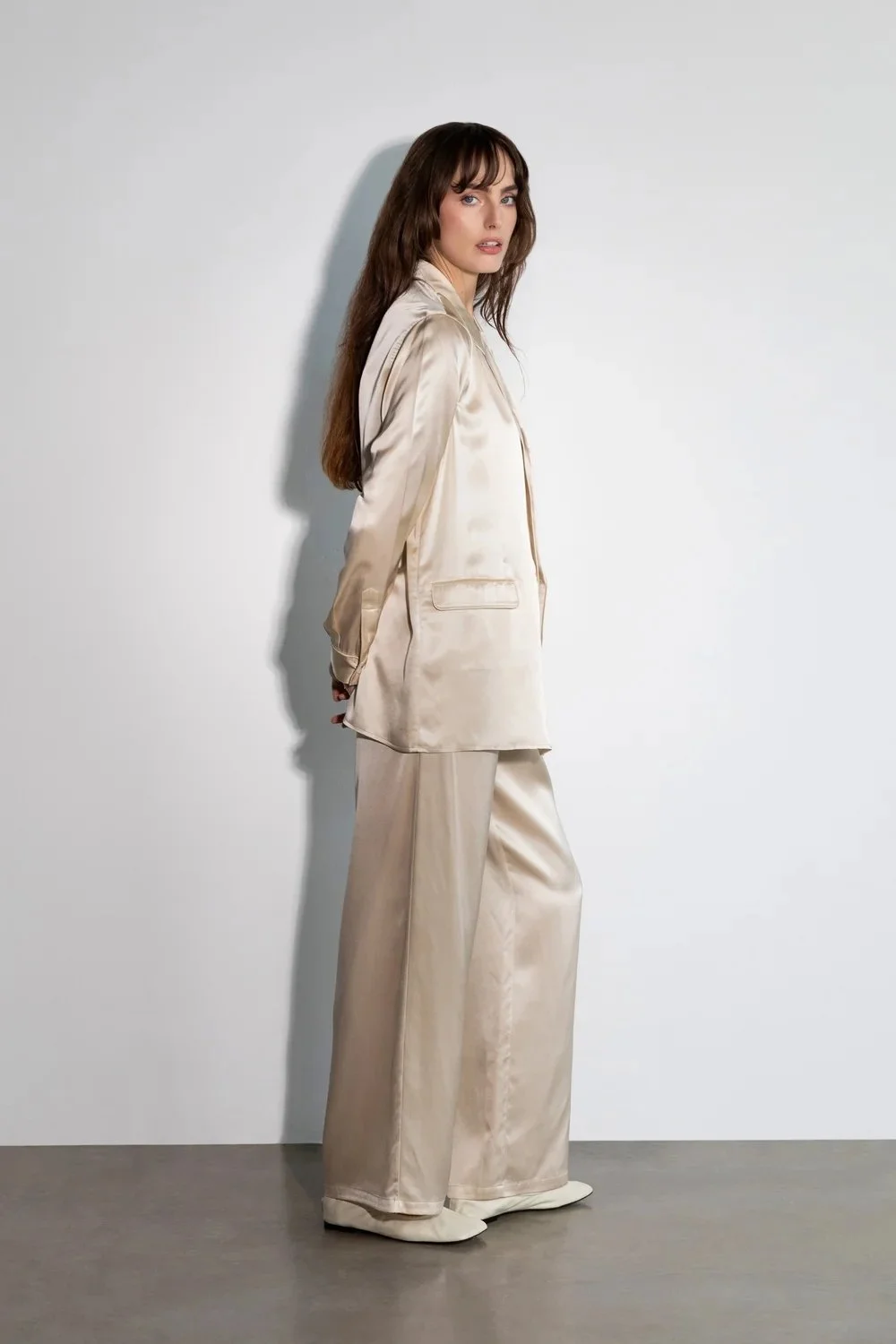A woman in beige silk pajama set standing against a plain white wall