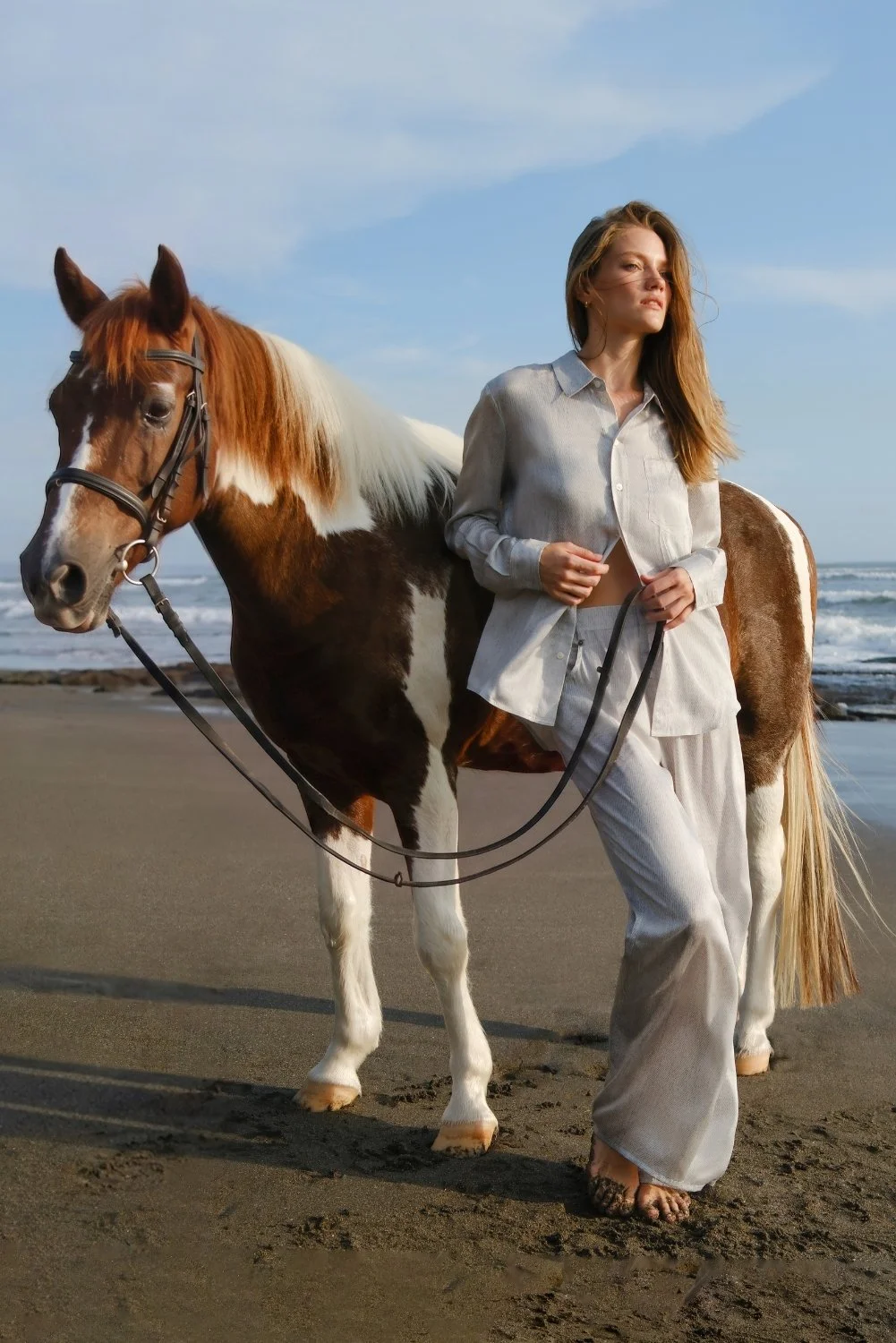A woman walking barefoot on the beach with a pinto horse, holding the reins. The woman is wearing light-colored pajamas and a button-up shirt, with the ocean and blue sky in the background.
