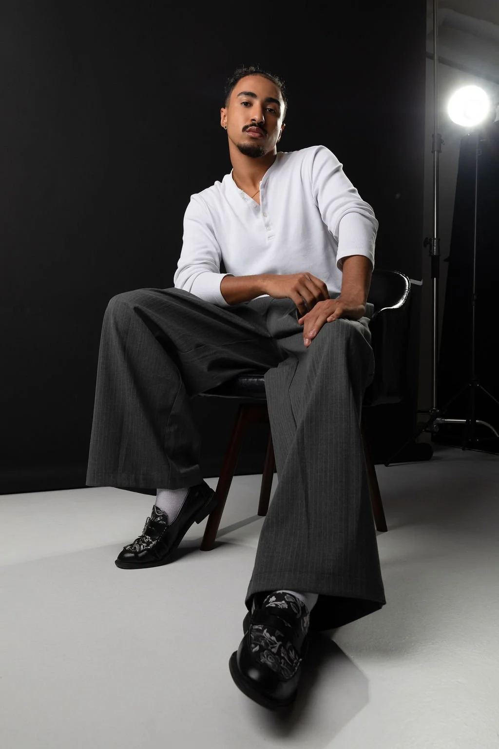 A man in a white long sleeve shirt and gray plaid pants sitting on a black chair in a photography studio with a dark background and studio lights.