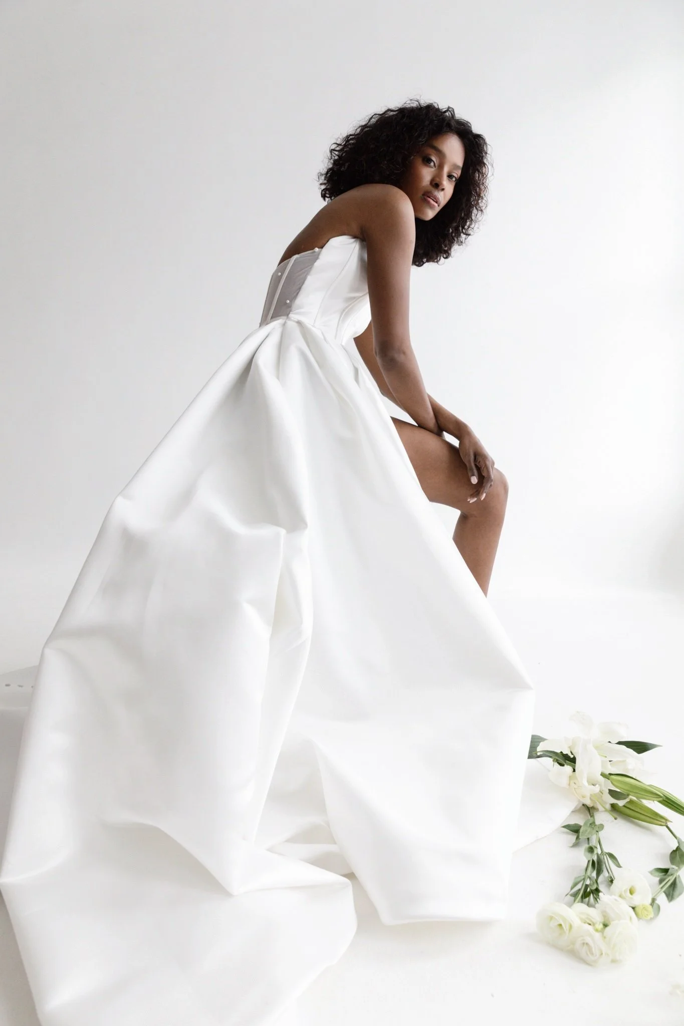 A woman with curly black hair is sitting on a white platform, wearing a strapless white gown with a full skirt. There are white flowers and greenery on the floor next to her, and the background is plain white.