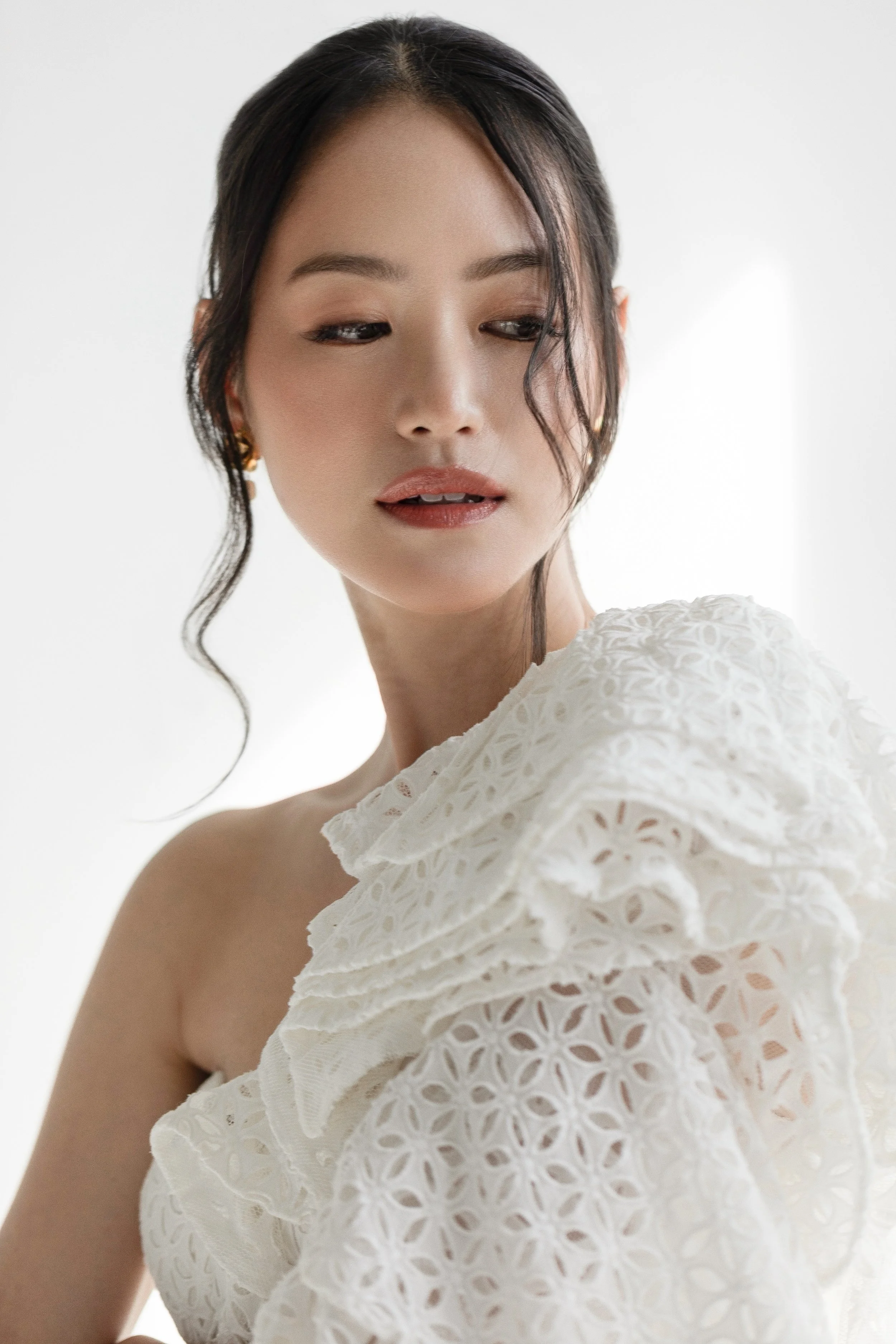 A woman with dark hair styled with loose curls, wearing gold earrings, and a white eyelet dress with ruffled details, facing slightly to her left against a white background.